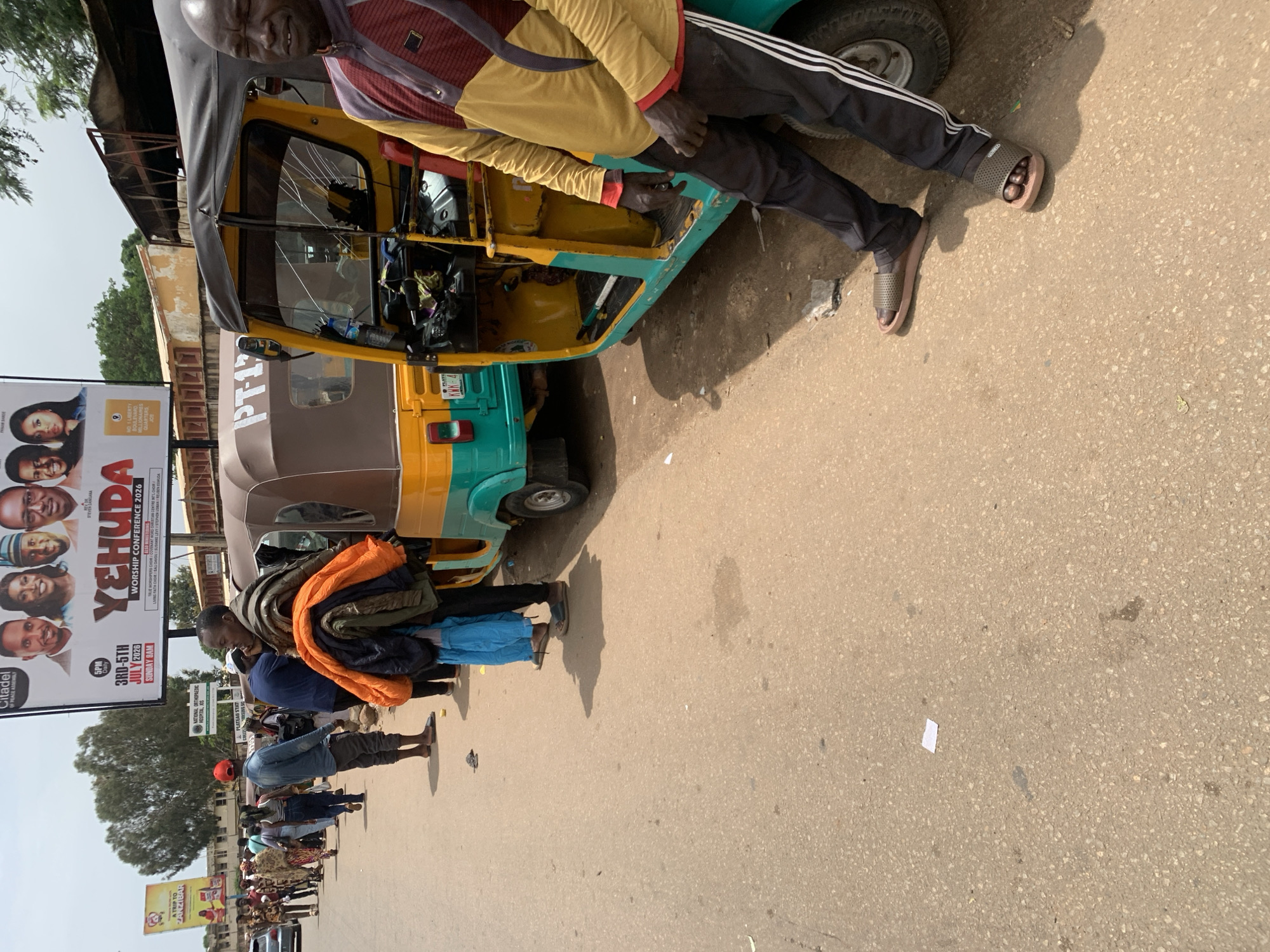 The image depicts a street scene in Jos, Nigeria, characterized by a row of people standing alongside parked auto-rickshaws, commonly known as "Keke Napep." In the foreground, a man with dark skin, wearing a yellow and burgundy patterned shirt and black athletic pants with white stripes, leans against a teal and yellow auto-rickshaw. He is looking towards the right side of the frame with his eyes closed and mouth slightly open, his expression suggesting discomfort or weariness. His feet are clad in brown slide sandals.
Behind him, another auto-rickshaw, primarily brown with yellow and teal accents, is visible, with a collection of bundled clothing, including an orange jacket, resting on its side. Several individuals are standing behind this auto-rickshaw, facing left. They appear to be a group of people, some of whom are carrying belongings.
Further down the street, a line of people are walking or standing, and more auto-rickshaws are parked. The environment appears to be an urban setting with a paved road and buildings visible in the background, including what appears to be a hospital or clinic sign that reads "NATIONAL ORTHOPAEDIC HOSPITAL, ENUGU." A large billboard with portraits of several individuals and text related to a "YEHUDA WORSHIP CONFERENCE 2026" is also prominent in the background, suggesting a public event or religious gathering. The sky is overcast, indicating a cloudy day. The overall scene conveys a sense of everyday life and transit in a Nigerian city.