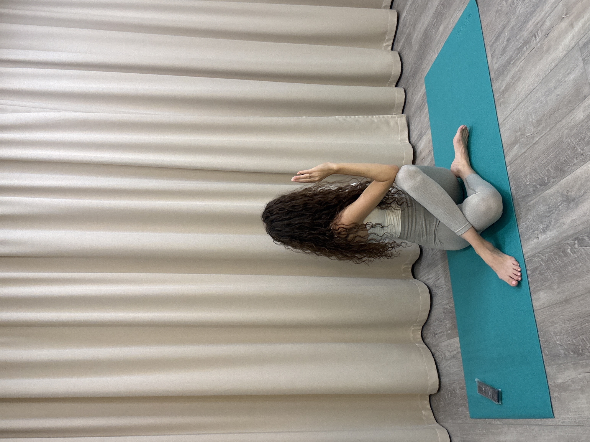 A person with curly brown hair is performing a yoga pose on a turquoise yoga mat. They are wearing a light-colored tank top and gray leggings. Their body is twisted to the right, with their left leg bent and their right leg extended. Their right arm is bent at the elbow and their hand is raised with fingers extended. The yoga mat is on a wooden floor. Behind the person is a light-colored curtain. A black object, possibly a remote control, is on the left side of the yoga mat. The scene appears to be indoors, in a room with neutral lighting. It is not possible to determine the specific location in Khimki, Russia from the image alone.