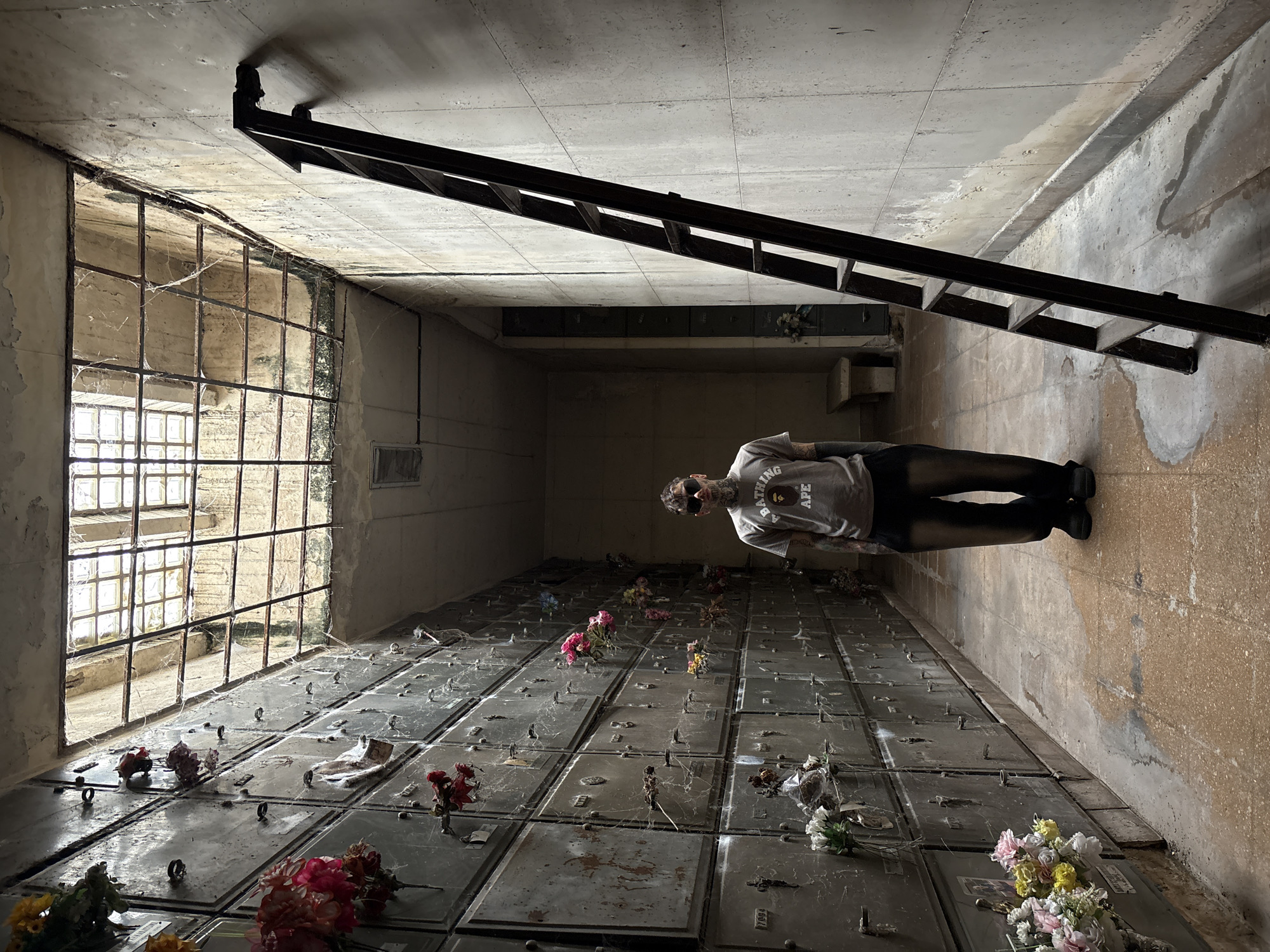 The image displays a solitary male subject positioned centrally within a narrow, aged columbarium corridor inside Recoleta Cemetery in Buenos Aires, Argentina. The subject, an adult male, presents with extensive tattoos covering his face, neck, and arms. He wears dark sunglasses, a light grey t-shirt bearing the "A BATHING APE" logo and text, dark baggy trousers, and dark footwear, standing upright and facing the camera directly.
To the subject's left, a tall wall of stacked rectangular crypt niches extends into the background. These metal or stone niches, many appearing weathered, are sparsely adorned with artificial flower arrangements and are partially obscured by numerous cobwebs. To the subject's right, a dark-colored, multi-rung ladder leans against a light-toned concrete wall, which exhibits prominent water stains and discoloration.
The ceiling above features a large, rectangular skylight covered by a metal grid, through which natural light enters. The skylight structure and surrounding ceiling show signs of rust, accumulated dirt, and extensive cobwebs. The tiled floor below is discolored, stained, and shows evidence of moisture. In the distant background on the right, a simple stone bench is partially visible. The entire environment indicates significant age, wear, and a degree of neglect.