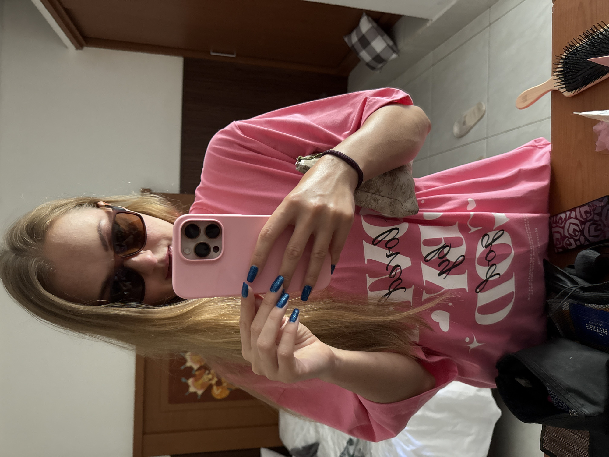 A young Caucasian female with long blonde hair, wearing dark sunglasses and a pink t-shirt with white text and heart graphics, is taking a selfie in a mirror. She is holding a pink smartphone in her right hand and a small, light-colored clutch bag in her left. Her nails are painted a sparkly blue. The background appears to be a hotel room in Denpasar, Indonesia, with a wooden wardrobe, a bed with white linens, and tiled flooring visible. A black toiletry bag and other items are on a wooden surface in the foreground.