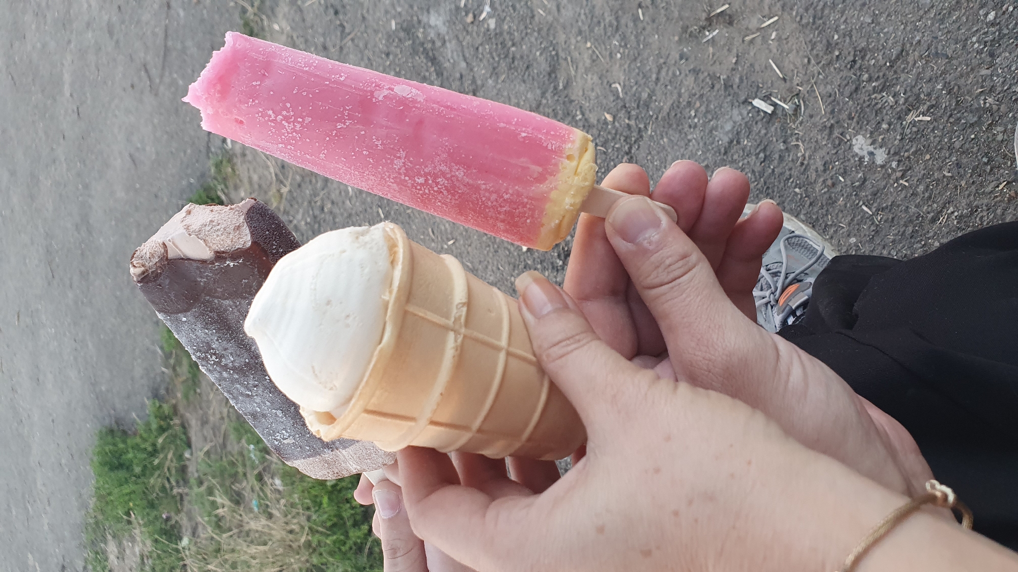 Three ice cream treats are being held by several hands in an outdoor setting. One is a chocolate-covered ice cream bar, partially bitten. Another is a vanilla ice cream cone, also partially consumed. The third is a pink popsicle, which appears to be strawberry or raspberry flavored with a lighter colored base. The ground in the background is a mix of dirt, gravel, and some sparse green grass. The image is taken from a close-up perspective, focusing on the hands and the ice cream.