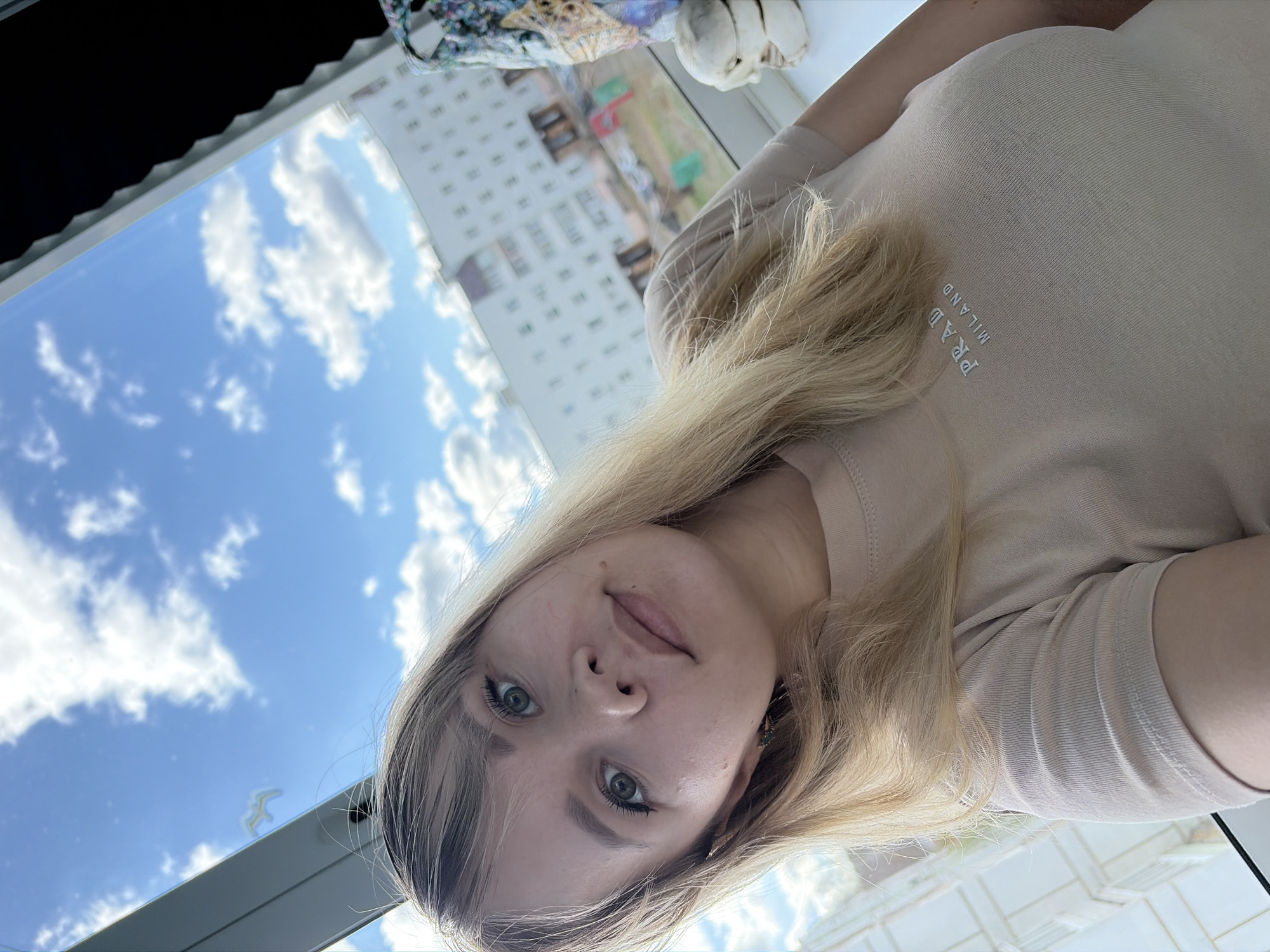 A rotated, selfie-style medium shot depicts a blonde-haired woman with gray eyes looking directly at the camera. She is wearing a beige "PRADA MILANO" shirt. The shot appears to be taken from an indoor location, with a bright blue sky visible through a window. A multi-story building is also visible through the window, its architecture implying an urban setting. The city and specific location are unidentified.