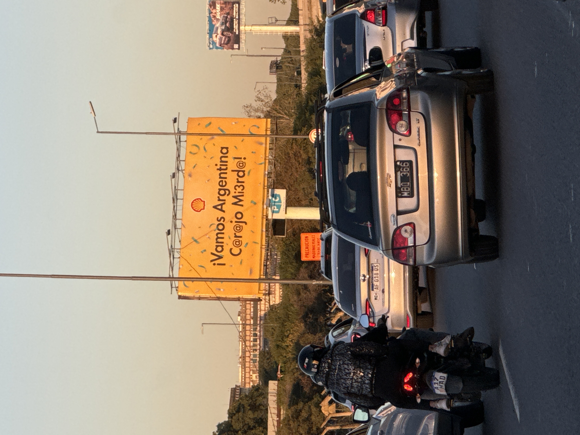 The image captures a scene of traffic congestion, likely in Buenos Aires, Argentina. Visible in the foreground is a motorcycle with license plate A17 HAD, ridden by a person clad in a black jacket and helmet. Cars are stalled on the road, their rear ends prominently displayed, including license plates such as MC AF 018 QS and MBQ 366. Dominating the background is a large, yellow billboard advertising Shell, with the message "¡Vamos Argentina C@r@jo Mi3rd@!" The billboard is partially obscured by street lighting and other infrastructure. Additional details include a cautionary sign warning of road machinery at work, and other billboards further in the distance. The environment suggests an urban setting with buildings and foliage lining the roadsides.