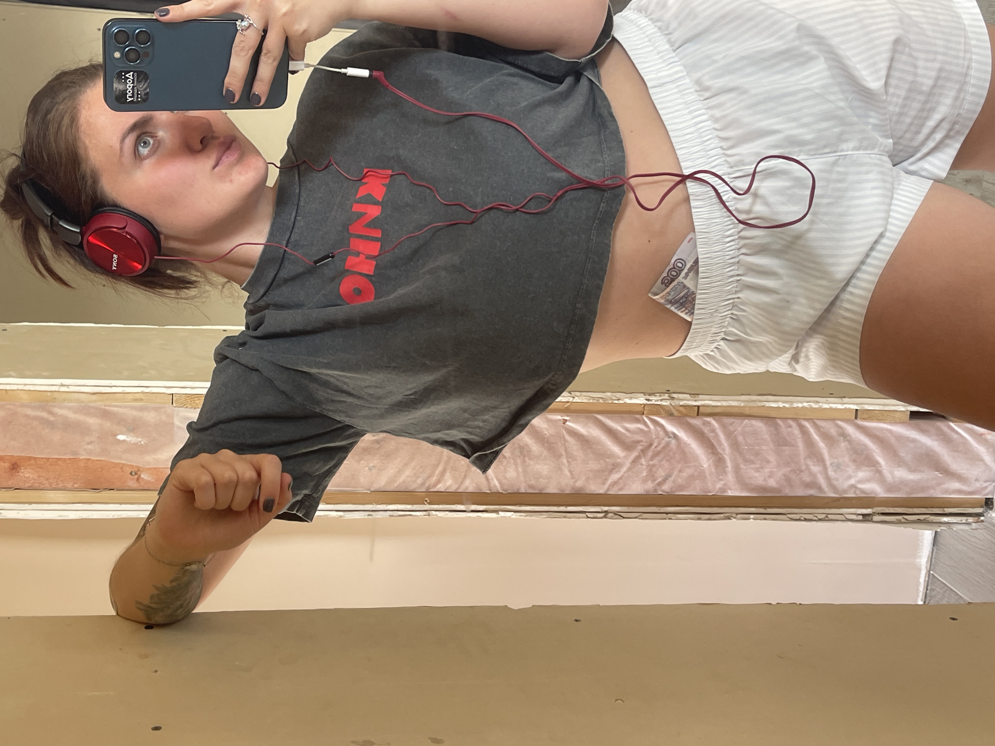 A woman is taking a selfie in front of what appears to be a renovation project. She has red headphones on and is wearing a grey cropped shirt and white striped shorts. A wad of cash is visible in her shorts pocket. Her right arm is holding up her body weight as she leans towards the camera. The background shows exposed wall framing covered in plastic. It's not possible to verify the specific location in Мосметаллоконструкция Онт, Russia from the image.