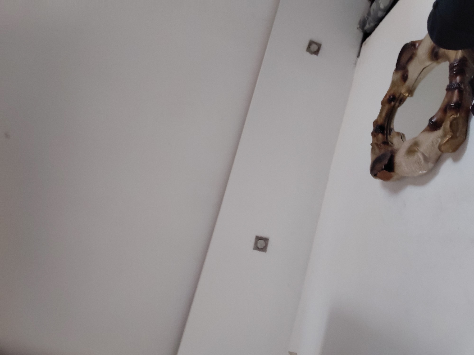 The image displays an indoor scene, characterized by light-colored walls and a ceiling, likely white or light gray, photographed from a low, upward angle. A prominent diagonal line separates these surfaces, creating a corner view of the room.
On the right side of the frame, a decorative, irregularly shaped mirror or wall art is partially visible. Its frame has an organic, gnarled appearance, composed of dark brown and beige sections that resemble natural wood, possibly bamboo, or a resin mimicking such material. The small, reflective surface of the mirror is visible within this intricate frame. At the very top right edge, a dark, soft object, possibly an article of clothing or fabric, is partially discernible.
Two square-shaped recessed fixtures, appearing to be lights, are embedded in the diagonal surface. These fixtures have dark centers within a lighter, metallic-looking square frame. The room appears brightly lit, likely by artificial light sources. The surfaces are plain and unadorned, contributing to a minimalist aesthetic. There are no people visible, nor any specific activities taking place. No text is present in the image. The time of day is not determinable, and no external weather conditions are evident.