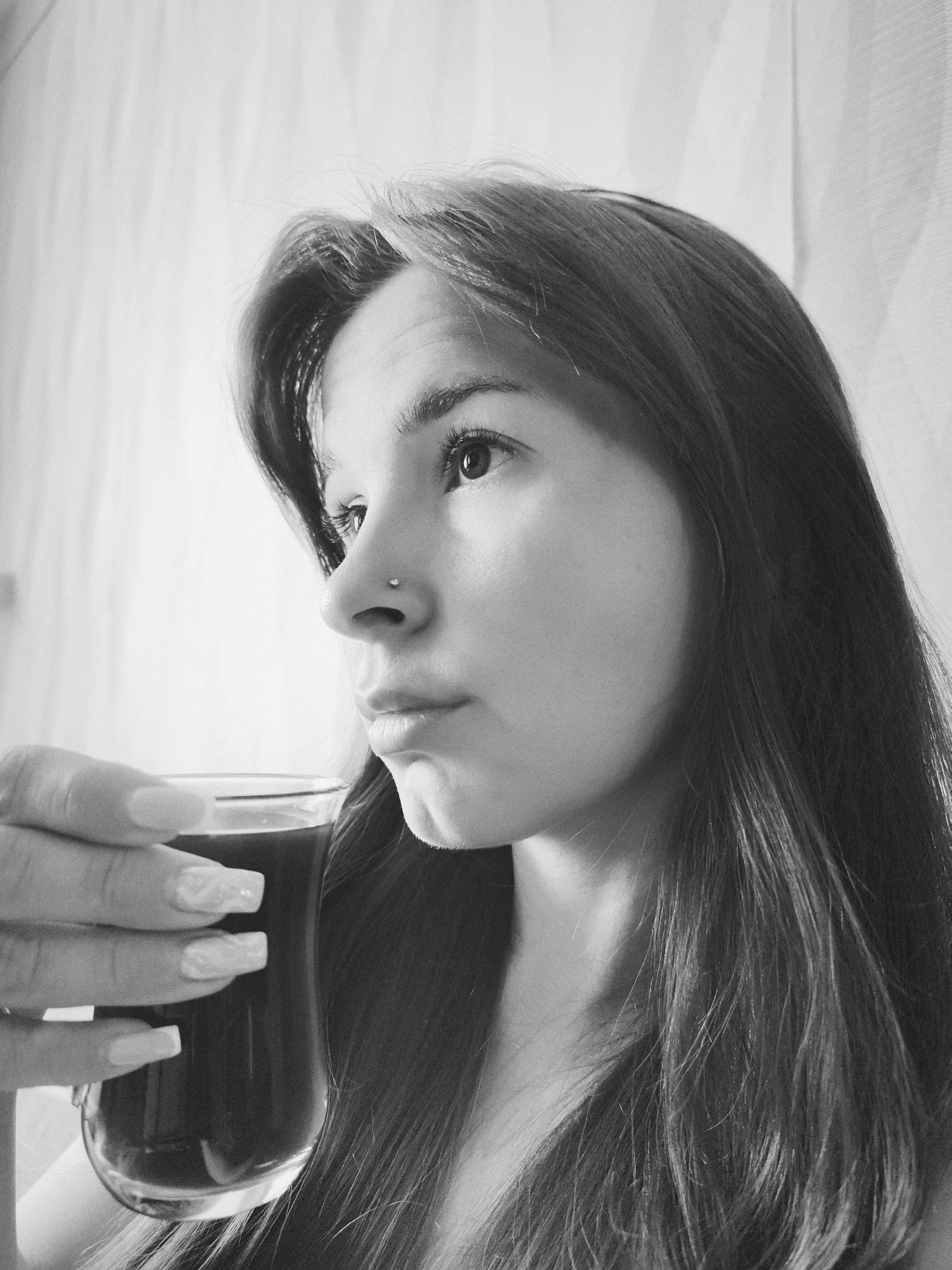 A black and white close-up photograph shows a woman holding a glass filled with a dark liquid. The woman is in the foreground, looking to the side with a contemplative expression. A small nose piercing is visible. Her long hair frames her face and falls down her shoulder. Her hand holding the glass is visible in the lower left, with long manicured nails. The background is a blurred, light-colored curtain. The photograph is taken indoors. There is no indication of location of the city of Фоки, Russia.