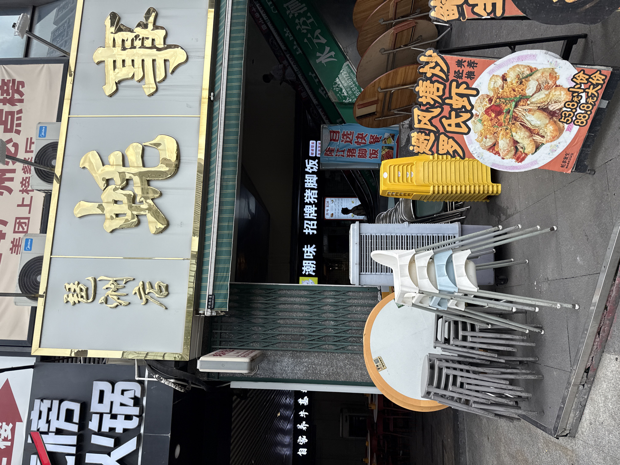 The image shows the exterior of a restaurant in Haizhu District, China. The restaurant's name, which appears to be written in gold Chinese characters, is prominently displayed on the facade. In front of the restaurant, several stacks of chairs are visible, along with a round table. A sign advertising a shrimp dish with prices is placed on an easel on the sidewalk. The entrance to the restaurant features an escalator leading to an upper level, with some patrons visible on the stairs. The overall scene suggests a busy commercial area.