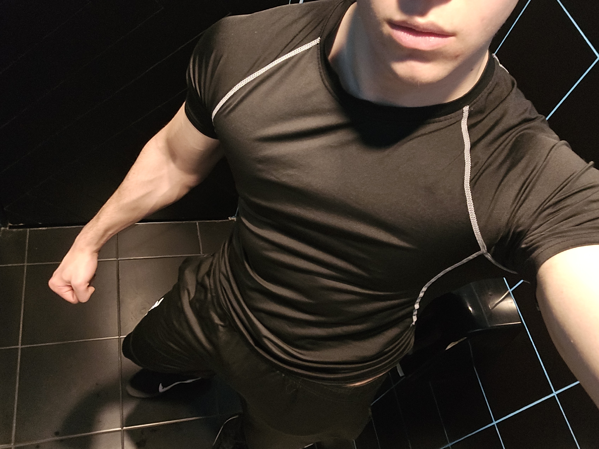An overhead-angle image depicts an unidentified male individual inside a building in Yerevan, Armenia. The subject occupies the majority of the frame, facing slightly away from the camera, which is positioned above and to the left of the subject. He is wearing a black, short-sleeved athletic or compression shirt with white stitching accenting the shoulder seams and sides, paired with black athletic shorts or pants. Black athletic shoes with a white Nike swoosh are visible on his feet. The subject's left arm is bent, with the hand clenched into a fist, positioned near his left hip. Only the lower portion of the subject's face, specifically the mouth and chin area, is visible. The environment consists of dark, likely black, square floor tiles with visible grout lines. The walls or background surfaces are also dark and feature faint, light-blue geometric lines, possibly reflections or architectural design elements on a reflective surface. The lighting is moderate, creating some shadows on the subject's clothing and the floor.