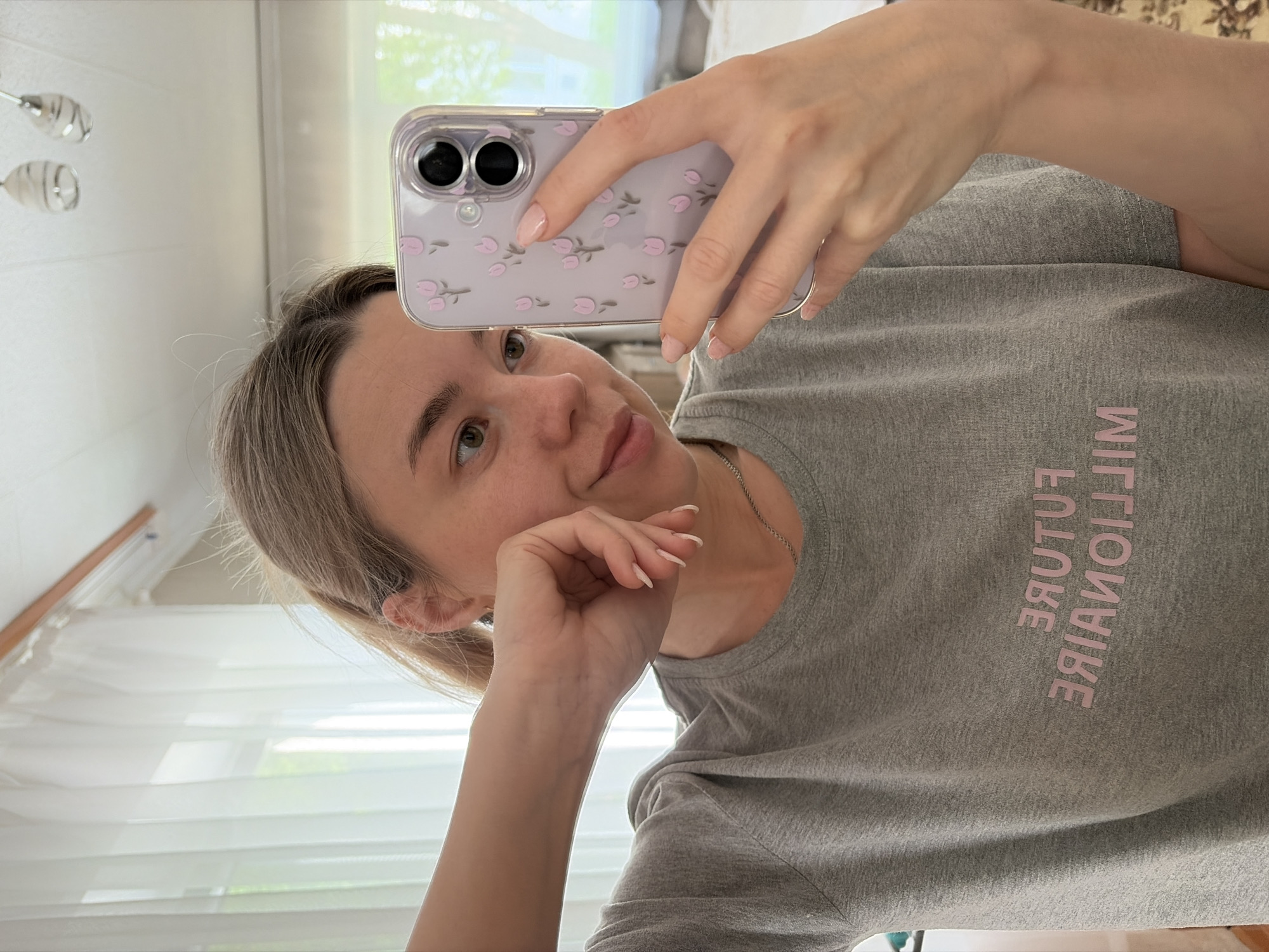 A woman with fair skin and light brown hair takes a selfie in what appears to be a brightly lit indoor space. She's wearing a gray t-shirt with the words "FUTURE MILLIONAIRE" printed in pink, facing backwards. Her right hand holds a mobile phone with a lavender floral case. Her left hand is near her face, resting on the chin. The background includes a partially visible window with light filtering through, a white blind, and a light fixture. The location of the image is unknown and is not apparent from the image. Therefore, the statement "location in the city of Chaikovsky, Russia" cannot be confirmed.