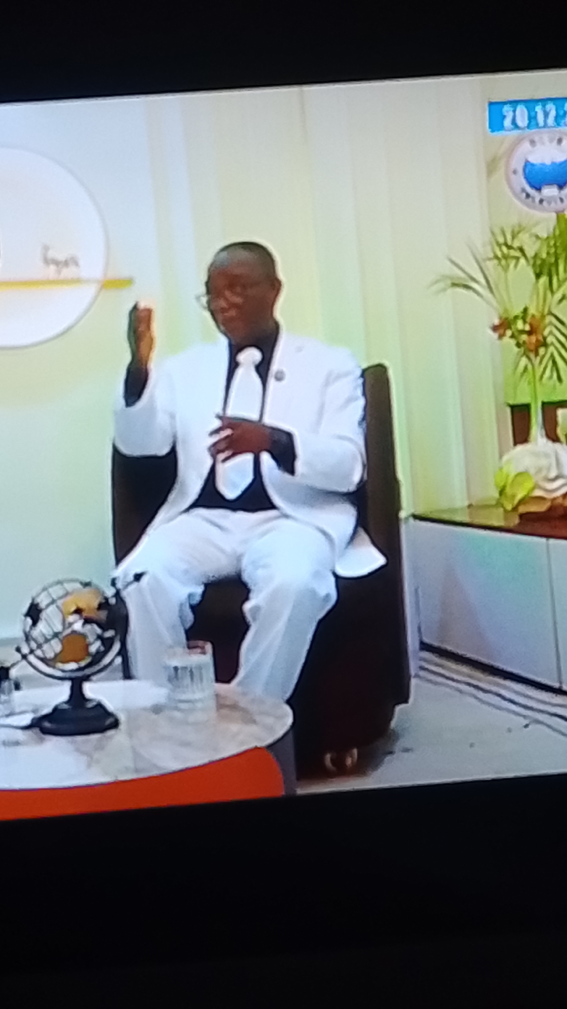 In Lokoja, Nigeria, a man dressed in a white suit, white shirt, and white tie is seated in a chair. He is wearing glasses and appears to be speaking or gesturing with his hands. To his left, on a table, is a globe and a glass of water. Behind him, a wall with a circular logo and a digital clock displaying "20.12.24" can be seen. The environment suggests a broadcast or presentation setting.