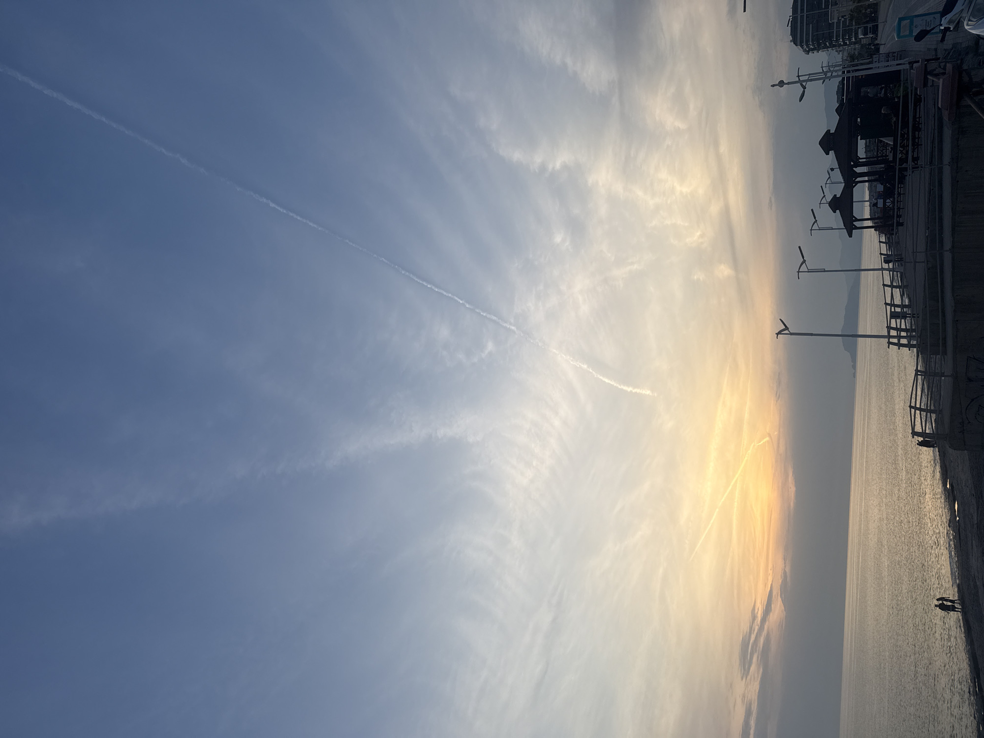 The image shows a waterfront scene with a view of the sky. The sky is predominantly blue with some white clouds and what appears to be a contrail from a plane. On the right side of the image, there is a cityscape with buildings, street lights, and some structures along the waterfront. The sea is visible below the cityscape. There are a few people discernible on the waterfront promenade. The overall environment suggests a serene or peaceful atmosphere.