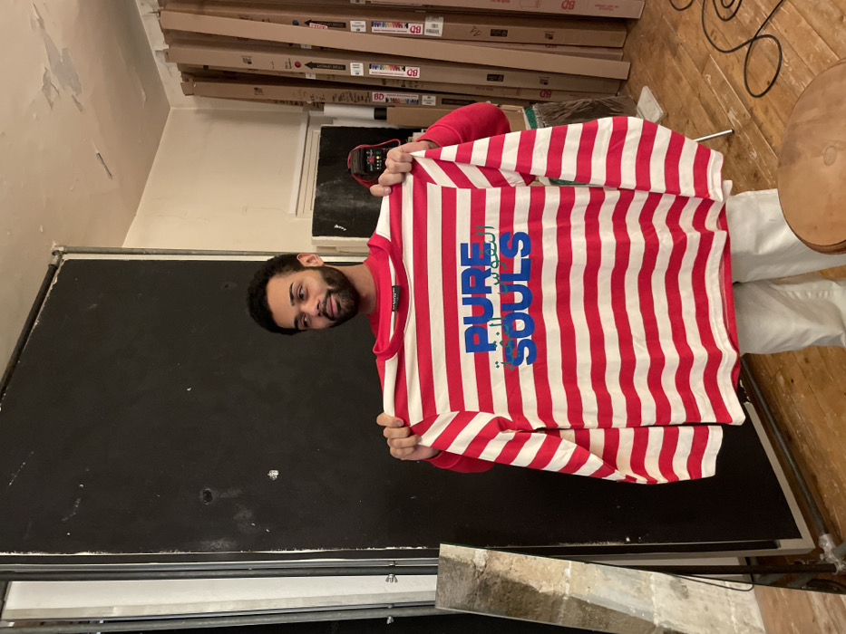 A man stands indoors, holding up a red and white striped long-sleeved shirt. The shirt has the words "PURE SOULS" in blue lettering with Arabic script. He is wearing white pants and a red shirt. Behind him are stacks of cardboard boxes and a black board with a metal frame. A wooden floor and table are visible on the right. The background also includes a wall with peeling paint.