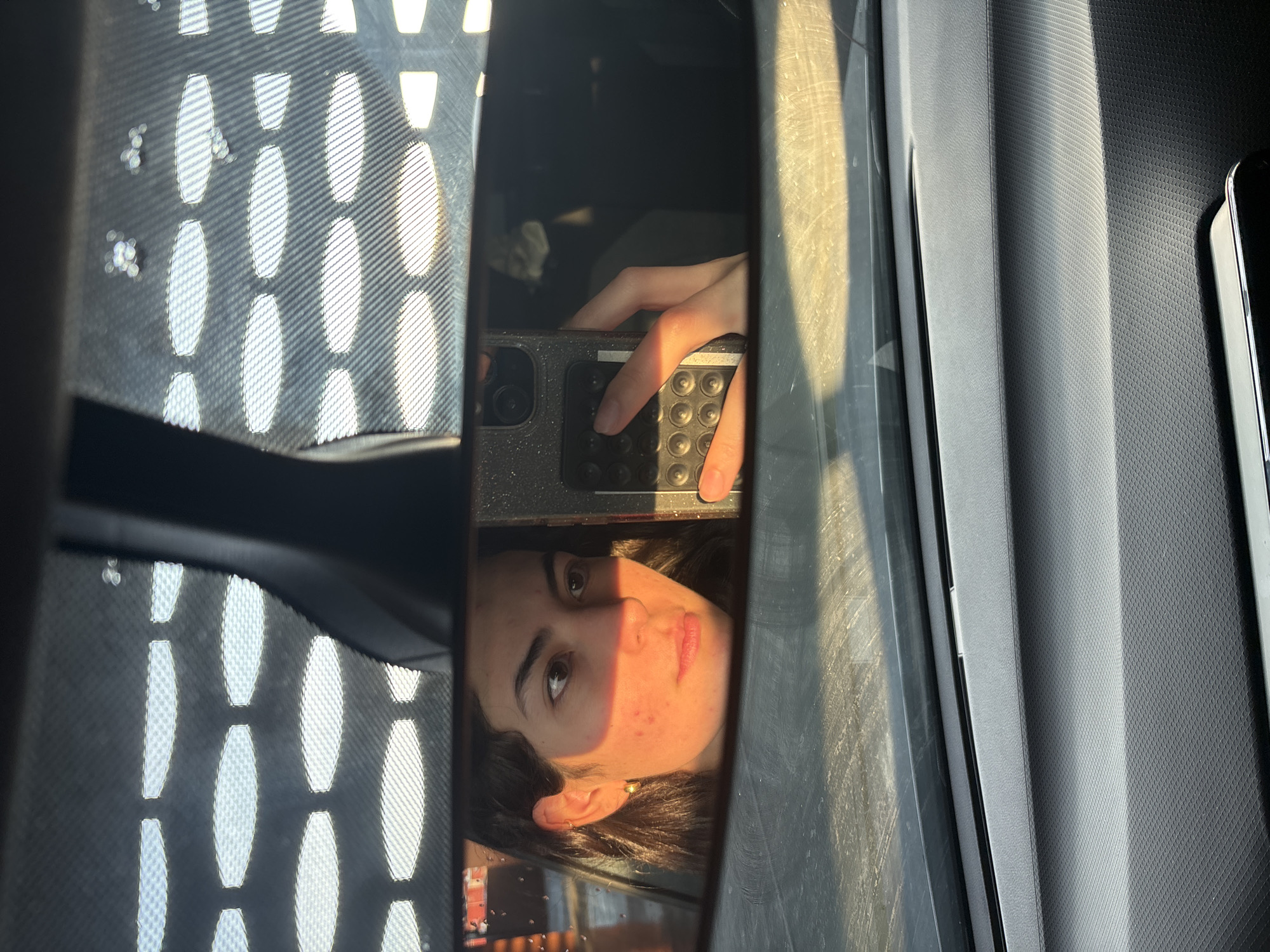 The image shows a selfie of a young woman reflected in what appears to be a car window or rearview mirror. She is holding a smartphone to take the picture, visible in the reflection. The background visible through the window has a pattern of elongated oval shapes, suggesting it might be a perforated sunshade or screen. The interior of the car has a textured gray surface in the background. There is insufficient information in the picture to verify that this photo was taken in San Bernardino Tlaxcalancingo, Mexico.