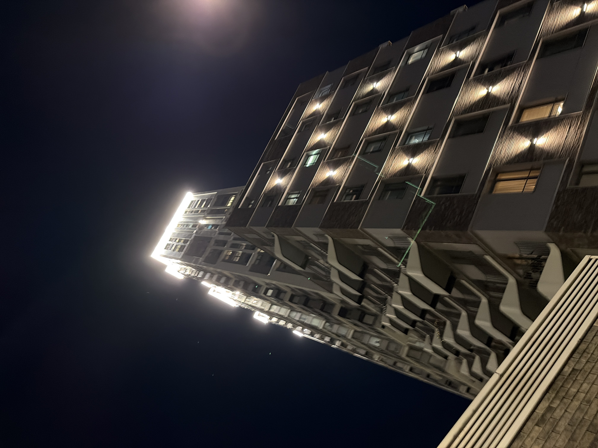 An upward, wide-angle shot captures a modern, multi-story apartment building at night. The building is illuminated by numerous lights, with particularly bright lighting around its upper sections and along the facade. Rows of windows are visible, some with lights on inside, indicating occupancy. The building's architecture features angular balconies and a textured brickwork pattern on certain sections. A streetlamp is visible to the right, its light casting a soft glow. In the foreground, a portion of a brick wall and a slatted barrier are present, suggesting a ground-level perspective or a nearby structure. The sky is dark, with a few faint specks that could be stars or distant lights. The overall impression is one of urban nighttime architecture.