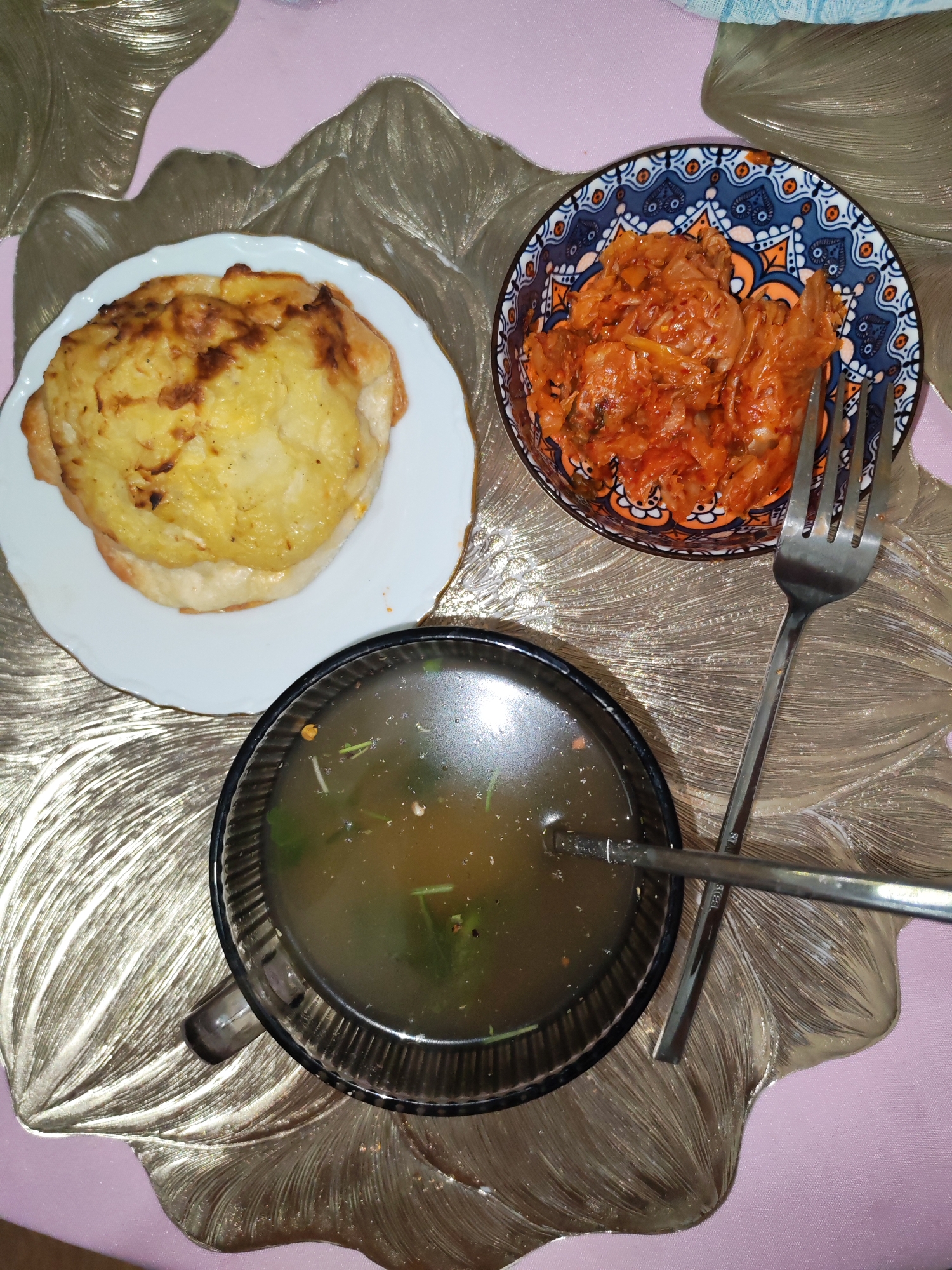 A top-down, close-up shot features a meal on a pink and gold placemat with a leaf design. The meal consists of three dishes: a round, golden-brown baked dish with melted cheese on top, served on a white plate; a small, decorative blue and orange bowl filled with red, spicy-looking food, likely kimchi; and a dark glass bowl containing a light brown broth with green herbs and small pieces of food. A silver fork is positioned in the kimchi bowl, and a spoon rests in the broth. The lighting is dim, casting soft shadows. The scene appears to be indoors, in a domestic setting, likely a dining table, in the city of Izhevsk, Russia.