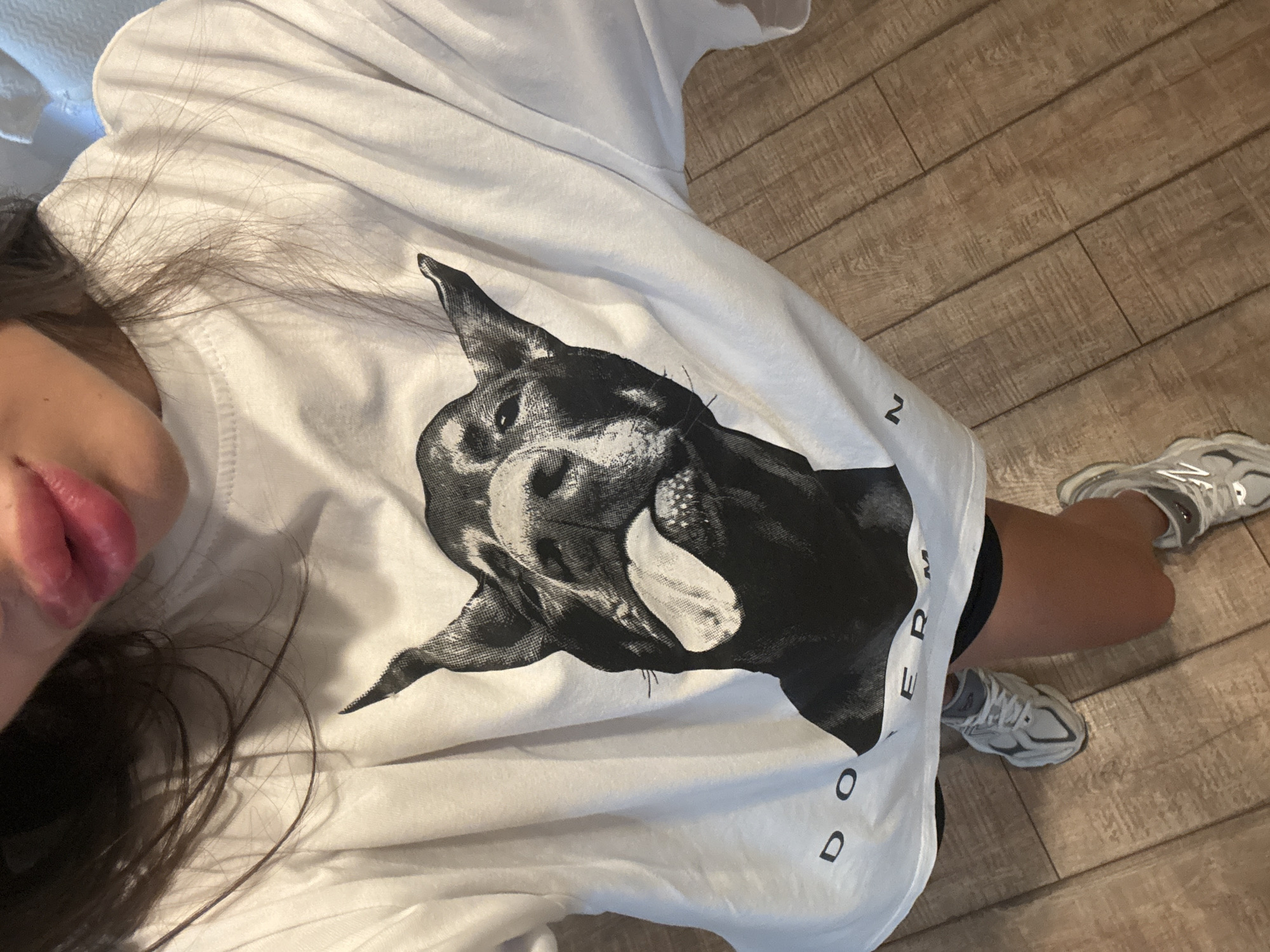 The image shows a close-up, overhead view of a person wearing a white t-shirt with a black and white graphic of a Doberman Pinscher sticking its tongue out. The t-shirt also has the word "DOBERMAN" printed on it. The person is wearing athletic shoes. The background consists of a wooden floor. The scene is indoors and there is no indication of the specific location within the city of Chișinău, Moldova, or any specific actions or interactions.