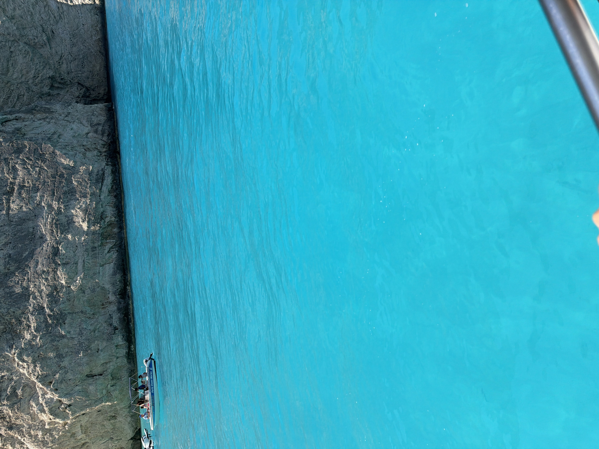 A small white motorboat filled with several people cruises on intensely turquoise waters near a sheer, rocky cliff face. The boat has a white canopy, and at least four individuals are visible, seated and standing. The cliff, a light gray and tan color, exhibits a rough, textured surface and a small cave-like opening near its base. The water is clear and appears shallow enough to see the sandy bottom in some areas. The scene suggests a sunny day with calm seas.