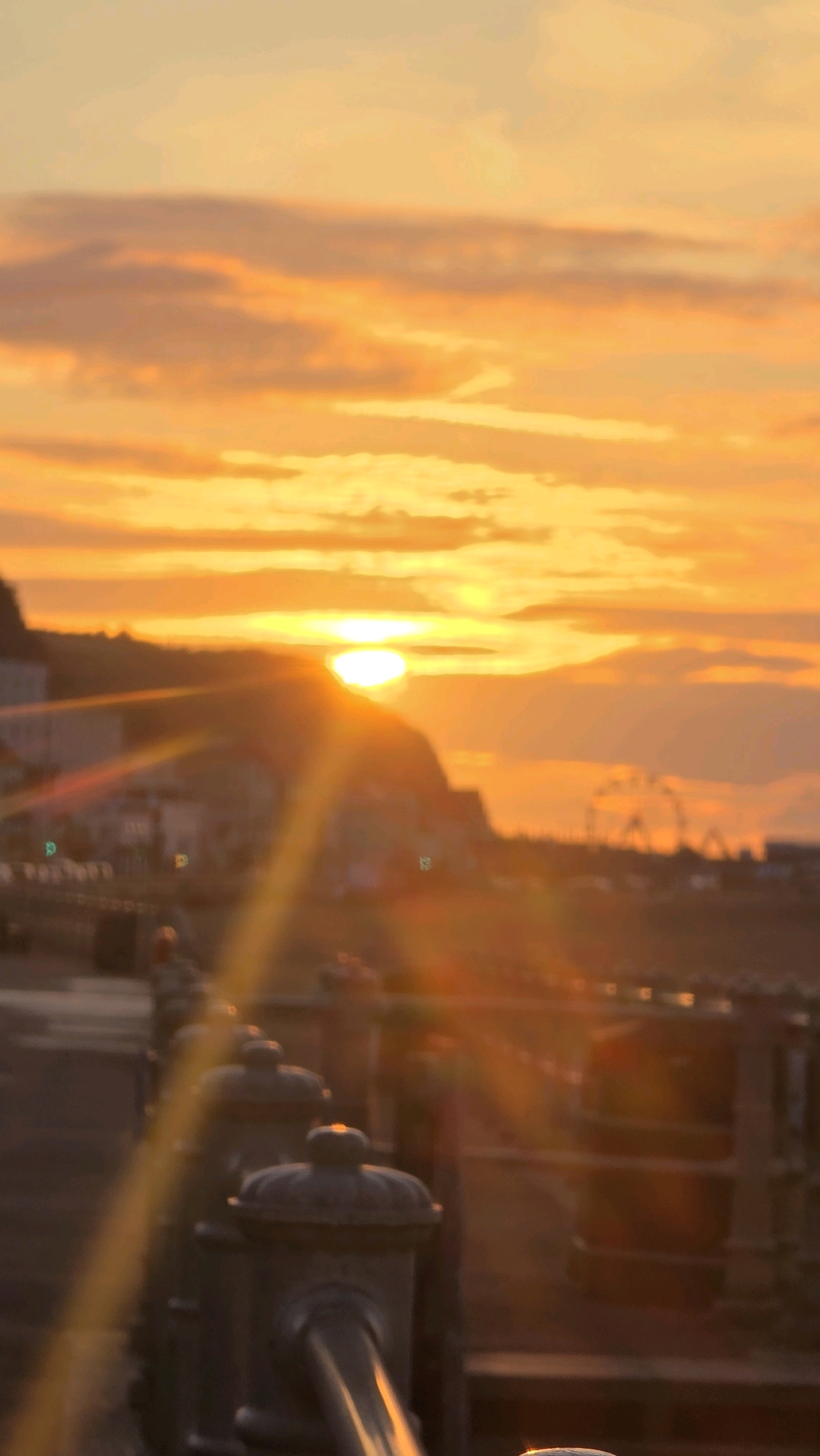 This is a photograph of a sunset over the coast of St Leonards, United Kingdom. The sun is low on the horizon, casting a warm orange glow across the sky and clouds. In the foreground, a metal railing with decorative posts lines a walkway. Beyond the railing, the blurred shapes of buildings and a Ferris wheel can be seen in the distance, suggesting a seaside town. The overall impression is one of a peaceful evening at the coast.