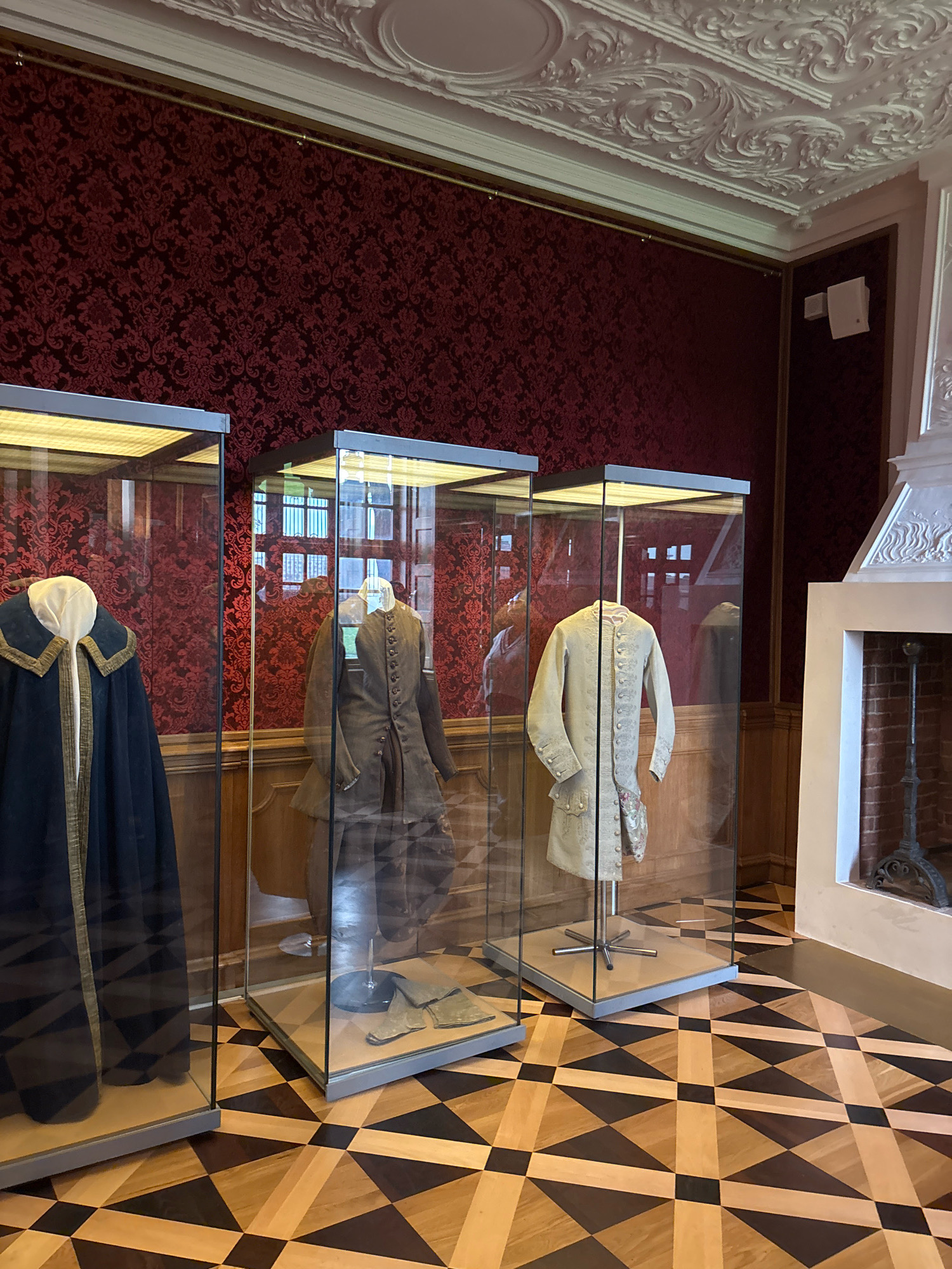 The image depicts a museum exhibit displaying historical garments in a room with ornate decor. The room features a red damask wallpaper with a floral pattern, elaborate white plasterwork on the ceiling and a fireplace surround, and a parquet floor with a geometric pattern of light and dark wood. Three glass display cases are arranged in the foreground, each containing a mannequin dressed in historical clothing.
The leftmost display case holds a dark blue cloak with gold trim, draped over a mannequin. The second case displays a brown, buttoned coat and a pair of gloves on a mannequin. The third case showcases a light-colored, ornate coat with detailed embroidery and buttons, and a small embroidered pouch. Reflections of windows and the opposite wall are visible in the glass cases. A decorative fireplace with a metal grate and tools stands to the right. The room appears to be well-lit, suggesting an indoor museum setting.
This exhibit is located in the Menshikov Palace in Saint Petersburg, Russia.