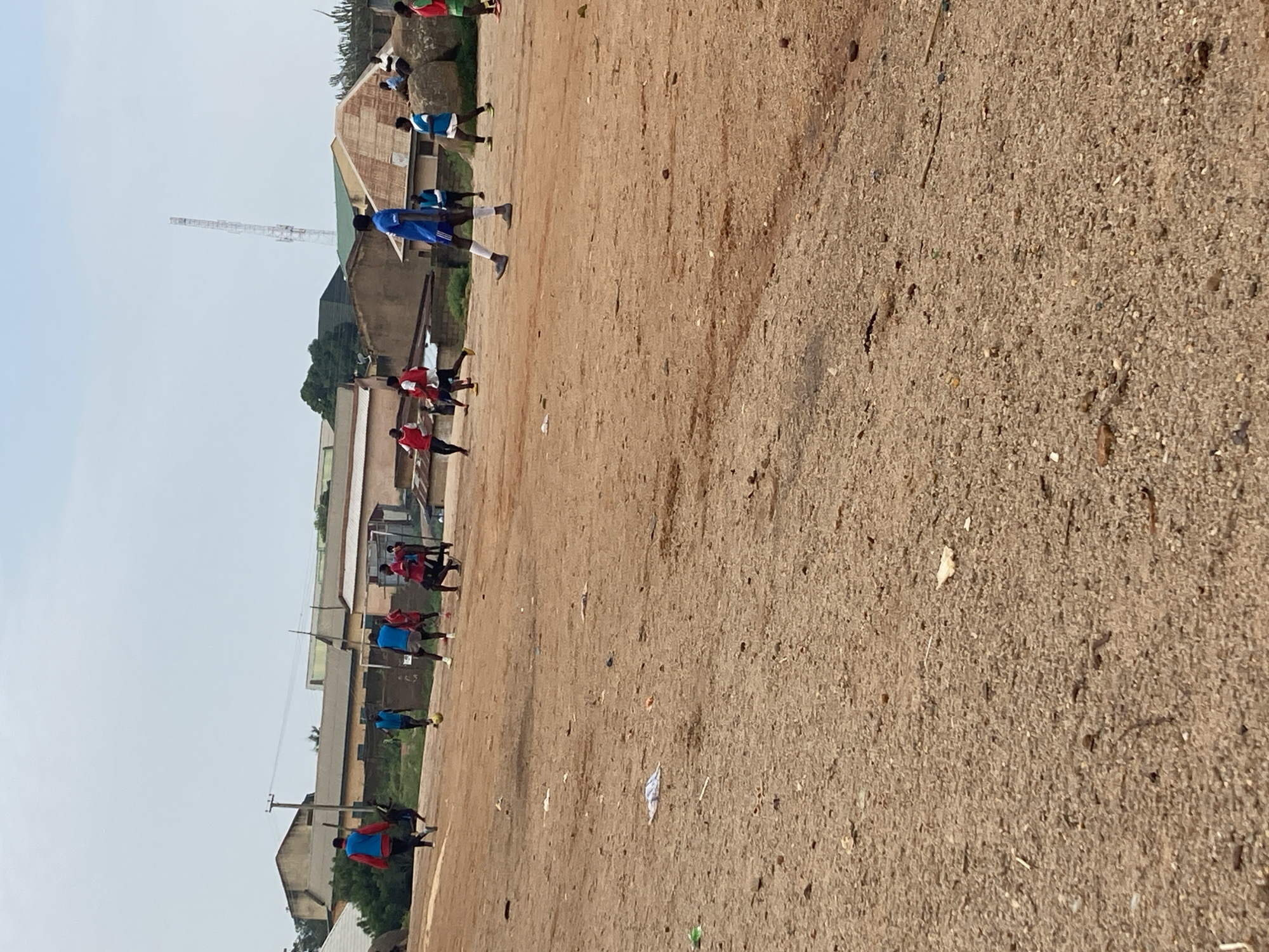 In an open, dusty field in Jos, Nigeria, a group of young men are engaged in a soccer game. The field is uneven and littered with debris, including scattered white paper and plastic. In the background, a cluster of buildings with red-tiled roofs and light-colored walls suggests a semi-urban or residential area. A tall cellular tower is visible in the distance. Several players are wearing athletic attire, with some in red and blue jerseys and shorts, while others are in blue jerseys and white shorts. One player in a blue jersey and white shorts is prominently walking towards the viewer in the foreground. The sky is overcast with a pale, cloudy atmosphere.