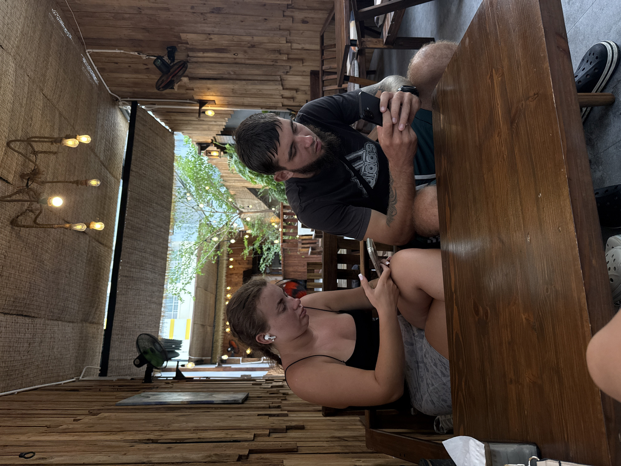 Two individuals, a woman and a man, are seated at a wooden table in an outdoor cafe with wooden walls and a thatched roof. The woman, on the left, has brown hair pulled back in a ponytail and is wearing a black camisole and a grey patterned skirt. She is looking down at her phone and has white wireless earbuds in her ears. The man, on the right, has dark hair and a beard and is wearing a black t-shirt with an Adidas logo and dark shorts. He is also looking down at his phone. The cafe appears to be open to the outdoors, with greenery and trees visible in the background. Lights are strung across the ceiling. The overall setting suggests a relaxed, casual atmosphere in Nha Trang, Vietnam.