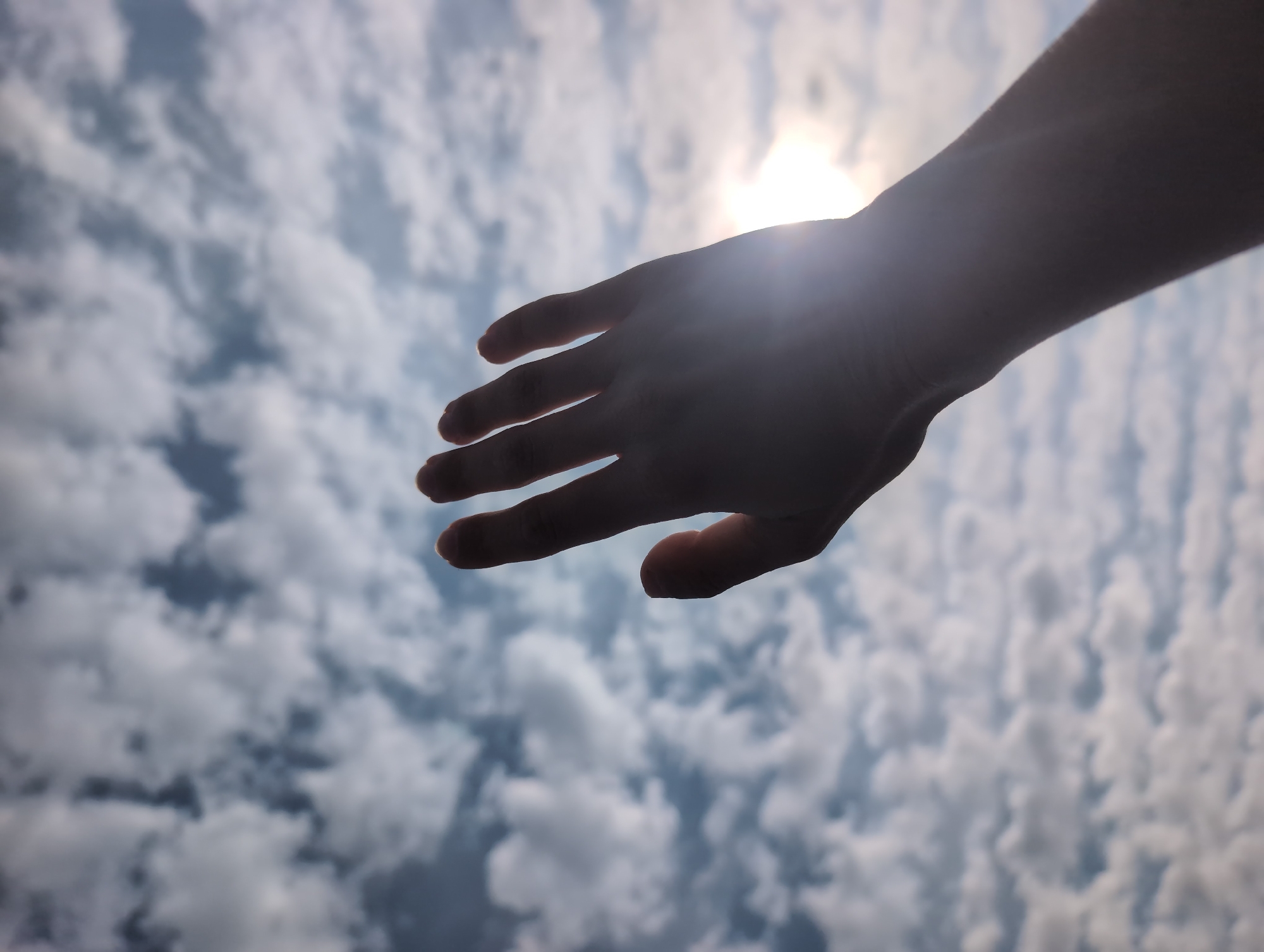 A hand is raised towards a cloudy sky, with the sun peeking through the clouds in the upper right corner of the image. The image is taken from a low angle, looking up. The clouds are white and fluffy, and the sky is a light blue. The hand is the only subject in the image. The image is of good quality with no visible artifacts. It is difficult to determine the exact location in Varengeville-sur-Mer, France from the image alone, but it is likely an outdoor location.