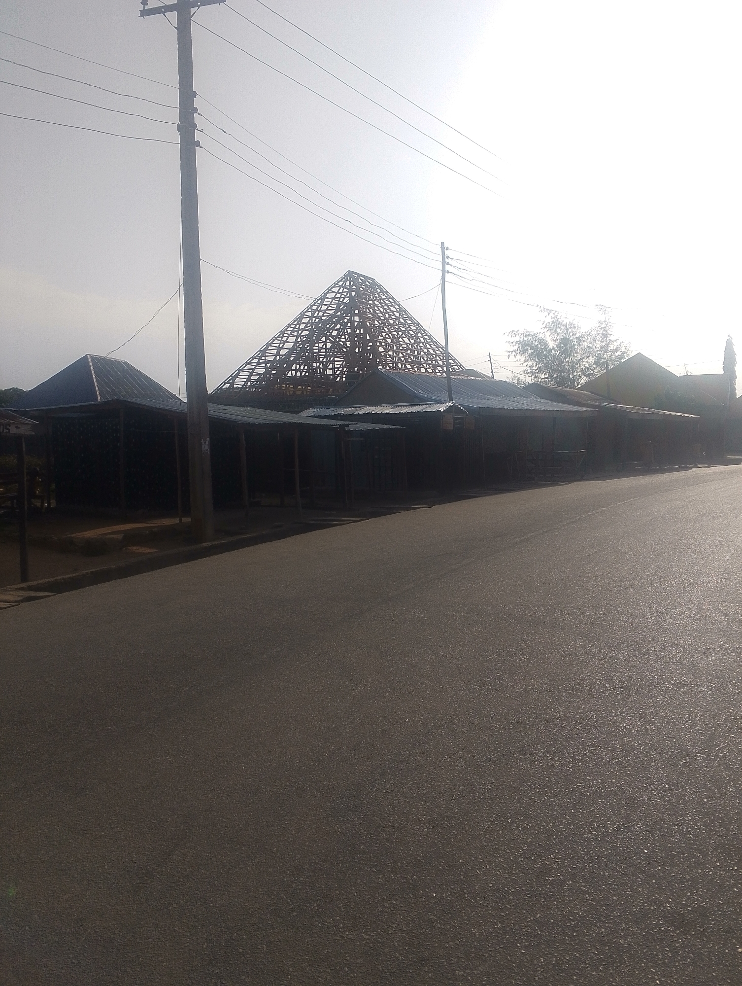 The image depicts a street scene in Jalingo, Nigeria, on a bright, hazy day. In the foreground, a paved road curves gently from the bottom right of the frame towards the left. Along the left side of the road, a row of market stalls with corrugated metal roofs and wooden supports are visible. One stall features a dark, patterned facade. Behind these stalls, the skeletal wooden framework of a large, pyramidal roof structure is under construction, suggesting ongoing development or renovation. A prominent wooden utility pole with multiple crossbars and overhead wires stands near the center of the image, casting a shadow on the road. In the background, trees and other structures, including a tall, slender building that might be a tower, are dimly visible through the bright, hazy sky, indicating a potentially arid or dusty atmospheric condition. There are no people visible in the image.