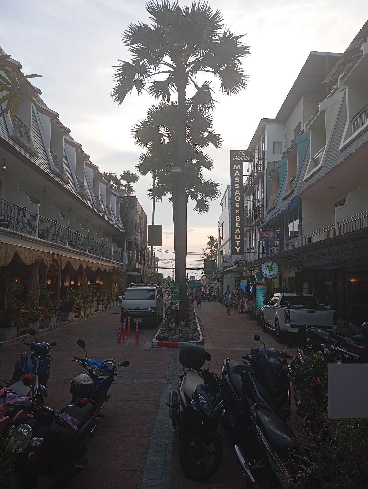The image depicts a street scene in Karon, Thailand, likely in the evening or early morning due to the soft lighting. A prominent palm tree stands in the center of the frame, with its fronds reaching towards the sky. The street is lined with multi-story buildings housing various businesses, including a "Natalie Massage & Beauty" establishment with a vertical sign. Several shops are visible on the ground floor, some with awnings and potted plants.
A silver van is parked on the right side of the street, along with several orange traffic cones. A white pickup truck is also parked further down the road. A row of parked motorcycles, predominantly scooters, occupies the foreground and the left side of the street. A few people are walking on the sidewalk on the right, with one woman in a patterned dress walking towards the viewer. The street surface appears to be paved with brick-like stones. The overall atmosphere suggests a commercial area with a tropical influence, typical of tourist destinations in Thailand.