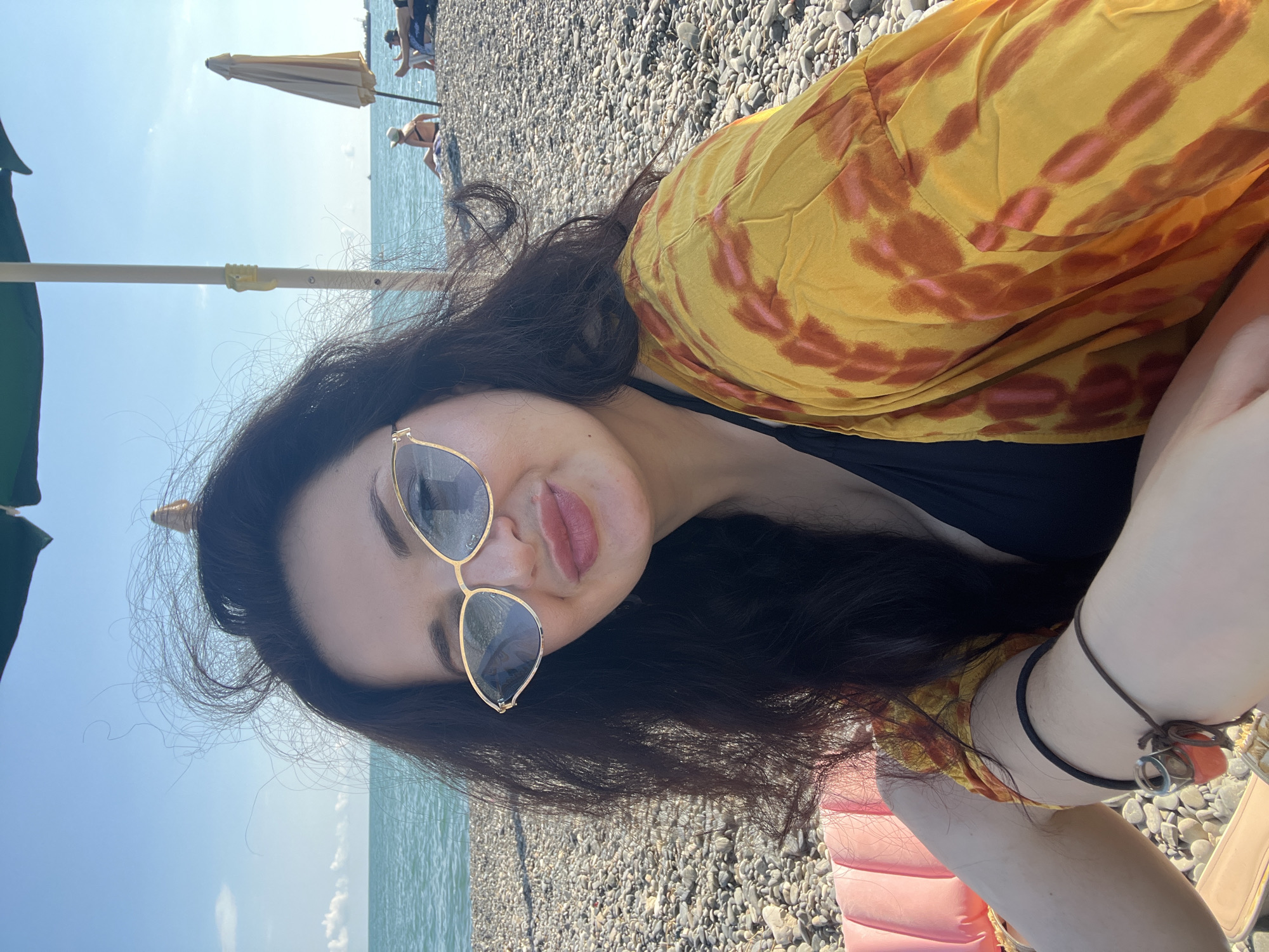 A medium shot selfie shows a woman with long, dark, wavy hair and sunglasses sitting on a pebble beach. She is wearing a black bikini top and a yellow and orange tie-dye cover-up. Her hair is blowing in the wind, with strands obscuring parts of her face and the background. The background shows the turquoise sea and a clear blue sky with a few wispy clouds. In the distance, on the beach, are other beachgoers, lounge chairs, and umbrellas. A green umbrella is visible directly overhead, with its pole to the right of the woman's head. A pink inflatable object is to her left, and a beach chair is partially visible below. The overall scene depicts a relaxed day at the beach in Sokhumi, Abkhazia.