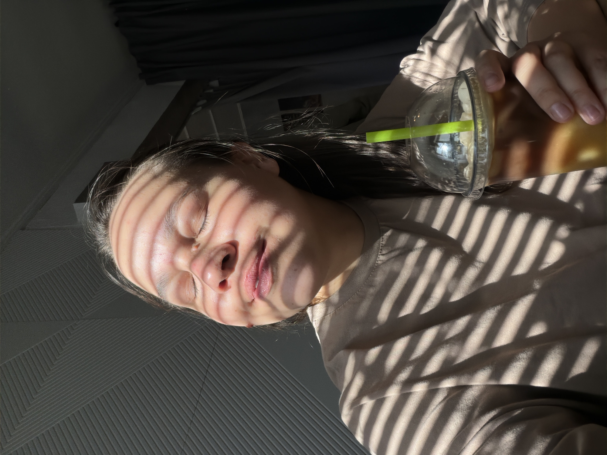 A person with closed eyes and shadow lines across their face and clothing holds a clear plastic cup with a green straw in their right hand. The cup appears to contain a blended beverage, possibly with whipped cream. The person is wearing a light brown or beige colored top. The background includes a gray textured wall on the left and a dark curtain on the top. The location is unknown, and there is no visible information to indicate the city of Chaikovsky, Russia.