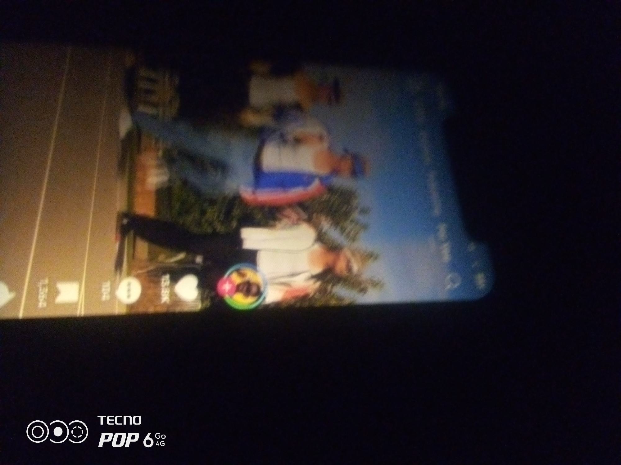 The image shows a close-up of a smartphone screen, displaying a video or social media feed. The video appears to feature two individuals, possibly men, standing in an outdoor environment. One person is wearing a white shirt and dark pants, while the other is in a blue and red jacket with white pants. They are positioned in front of a backdrop that includes greenery and a sky, suggesting it might be a park or a natural setting.
The phone's interface is partially visible, with icons and text elements indicating it is a digital platform. There are numbers such as "1,354", "104", and "15.8K" displayed, likely representing engagement metrics like likes or views. A circular icon with multiple colored segments is also present, possibly a profile picture or a feature icon. The bottom of the screen prominently displays "TECNO POP 6 Go 4G", indicating the brand and model of the phone. The lighting and overall quality of the image suggest it was taken in low light conditions or with a lower quality camera.
There is no visible information within the image or its crops that can be used to determine the specific location in the city of Howayi, Nigeria.