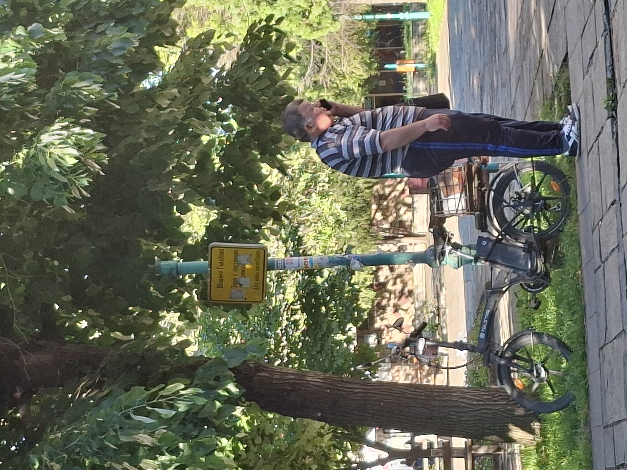 An older man is standing on a sidewalk in Galabovo, Bulgaria, holding a cell phone to his ear. He is wearing a striped shirt, dark pants with a blue stripe down the side, and blue and white shoes. He is standing next to a black electric bicycle parked on the sidewalk. A yellow sign is attached to a green pole near the bicycle. The sidewalk is paved with gray stones, and there is grass to the right of the sidewalk. A tree with green leaves is in the background to the left, and there are buildings in the background.