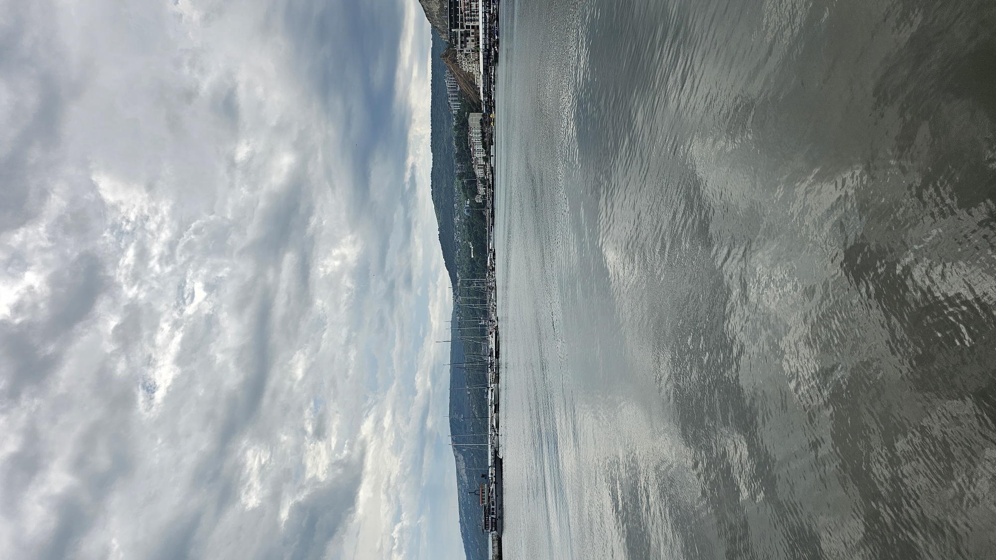 The image depicts a harbor scene in Balchik, Bulgaria, under an overcast sky. The upper half of the frame is dominated by a dense layer of light and dark gray clouds, with brighter patches indicating thinner areas. The lower half features calm, rippled water, reflecting the cloudy sky in muted tones of gray.
Along the left side of the harbor, a long, multi-decked vessel, possibly a tour boat or ferry, is docked. To its right, numerous sailboats with tall, slender masts are moored in a line. Behind these boats and extending across the mid-ground are rolling green hills covered in lush vegetation, with various buildings and structures interspersed along their slopes. In the far background on the left, a light-colored cliff face is visible.
Towards the right side of the frame, several multi-story buildings, potentially part of a hotel or resort complex, are constructed into a steep, rocky incline overlooking the water. More structures and what appear to be smaller piers or waterfront establishments are visible along the right edge of the harbor. There are no discernible people or active interactions evident in the scene. The overall atmosphere is serene and muted due to the cloudy conditions.