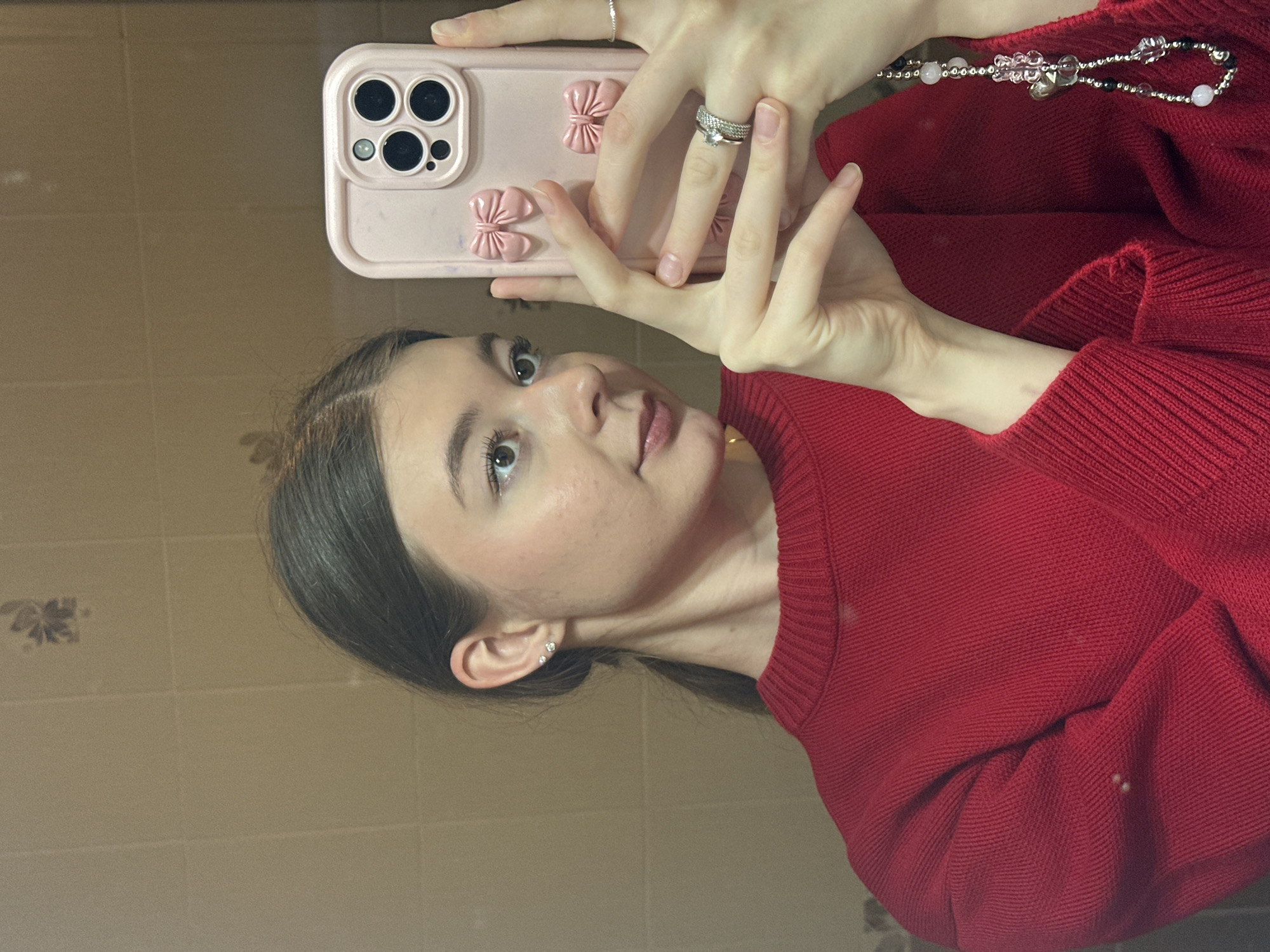 A mirror selfie captures a young woman, likely in Kazan, Russia, posing indoors. She is seen from the chest up, with her head slightly tilted, and her gaze directed towards the camera in her hand. Her dark brown hair is parted centrally and styled back from her face. She has light-colored eyes, subtle makeup with a pinkish lip gloss, and visible freckles across her nose and cheeks. She is wearing a red long-sleeved knitted or ribbed top.
In her right hand, she holds a light pink smartphone, which has a distinct camera bump and is encased in a light pink cover adorned with two prominent pink bow-shaped decorations. Her left hand is also visible, with fingers gently splayed around the phone. On her left ring finger, she wears two rings: a prominent silver ring with a clear stone and a simpler silver band beneath it. A thin silver band is also visible on her right pinky finger. Her left earlobe features two small, clear stud earrings. A multi-bead necklace, featuring white, pinkish, clear, and some black beads, along with a small bell-shaped charm, is partially visible around her neck, resting on her red top.
The background, reflected in the mirror, consists of light brown or beige tiled walls, suggesting an indoor setting like a bathroom or hallway. A faint, darker, possibly floral or abstract pattern is visible on one of the tiles. The lighting is soft and even, indicating indoor artificial light. There is no visible text in the image.