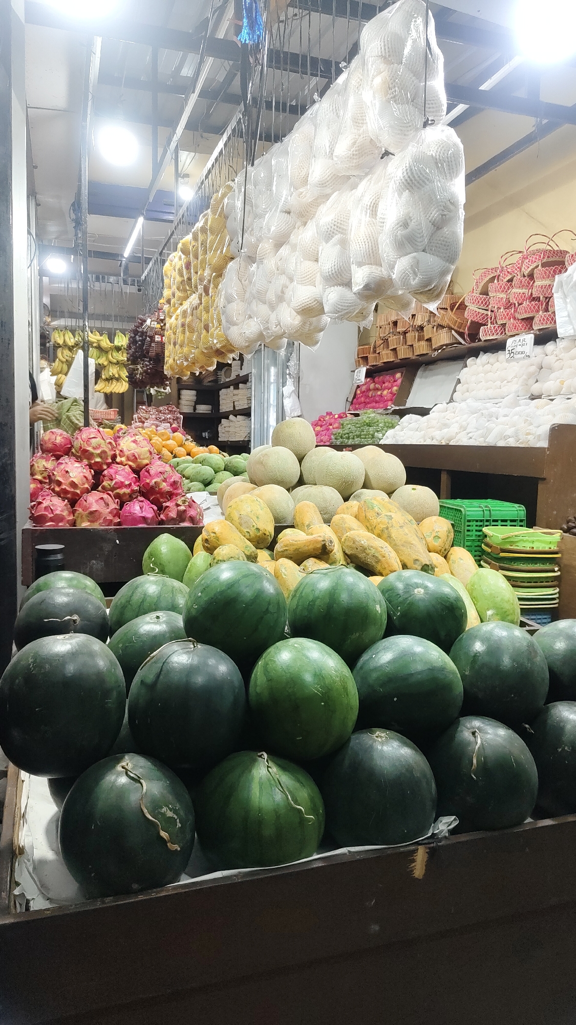 A fruit market stall in Malang, Indonesia displays a variety of produce. In the foreground, several large watermelons are neatly stacked, some with visible tendrils. Behind them, a pile of lighter green fruits, possibly mangoes, is visible. Further back, dragon fruits, characterized by their pink and yellow skin, are arranged in rows. A selection of cantaloupe and papaya fruits are also present. Hanging from the ceiling are strings of bananas and grapes. In the background, baskets of various goods, likely more produce or crafts, are stacked. The market appears to be well-lit, with overhead fluorescent lighting. The overall scene depicts a vibrant and abundant fruit market.
