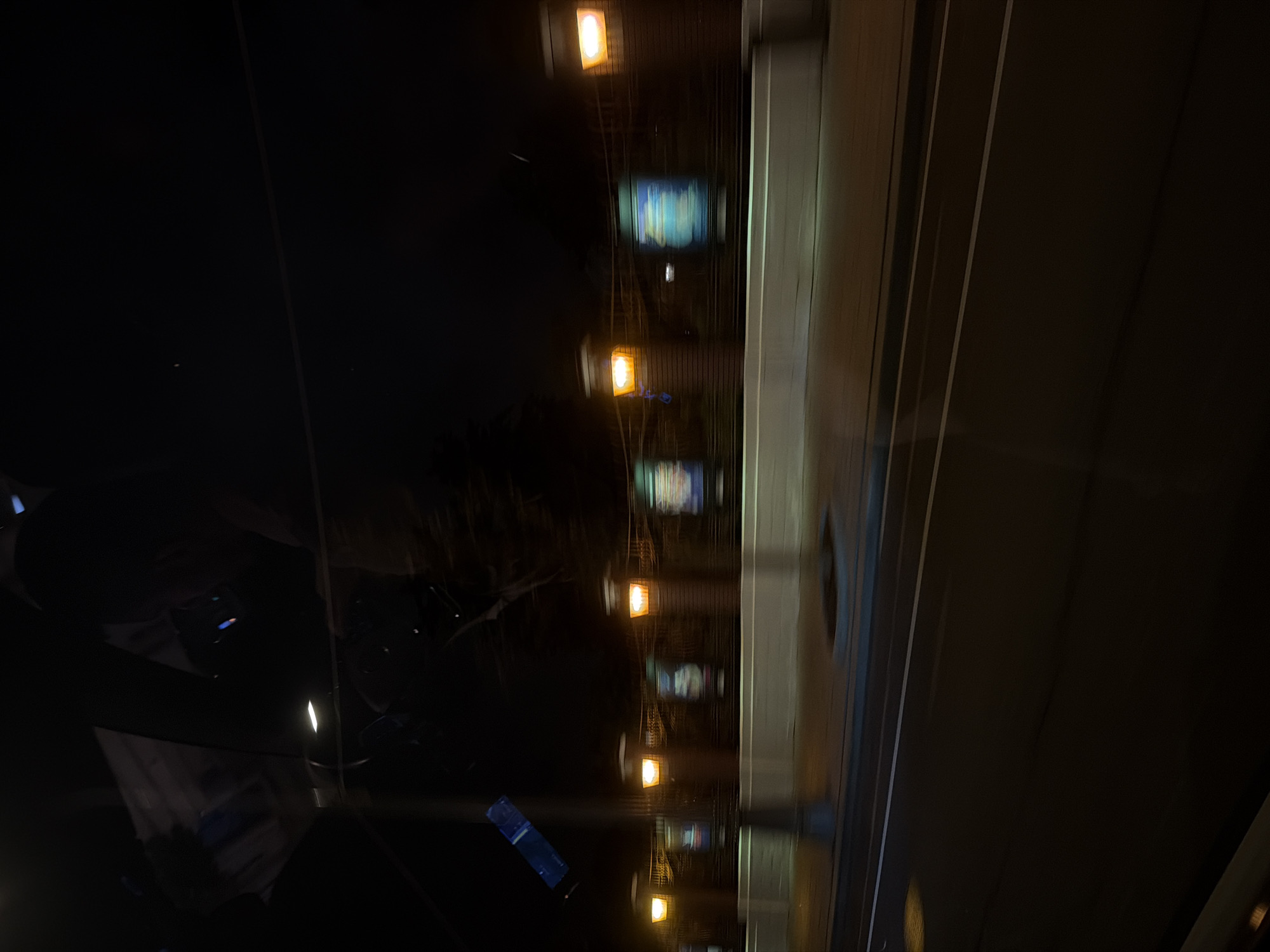 The image depicts a nighttime view of a dimly lit area, possibly a street, with multiple light sources arranged in a vertical pattern. The illumination is inconsistent, with some areas appearing darker than others. To the right of the image is a metallic panel with vertical lines. A blurred reflection can be seen on the left side. The location of the image is the city of Moscow, Russia.
