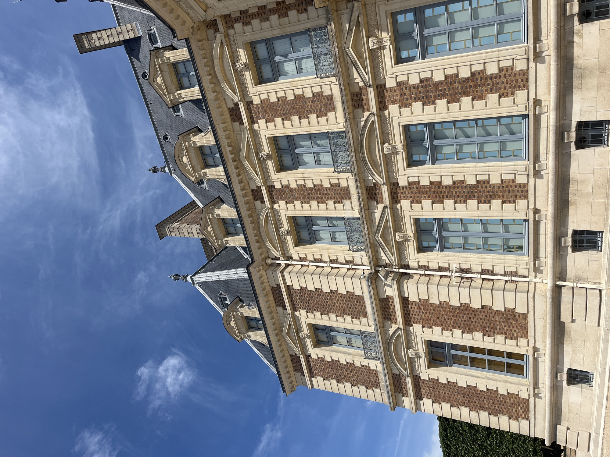 The image shows a close-up, upward-angle view of the facade of the Château de Sceaux, a historical building located in the Parc de Sceaux, in Sceaux, France. The architecture is characterized by its multi-story structure with a steeply pitched, dark grey slate roof featuring multiple dormer windows with decorative pediments. The facade itself is a combination of reddish-brown brickwork interspersed with decorative light-colored stone elements, including quoins, cornices, and window surrounds. The windows are large, tall, and divided into multiple panes with light blue frames. Some of the windows have ornate black wrought-iron balconies. The sky above is a vibrant blue with wispy white clouds. To the left, a portion of a meticulously maintained dark green hedge is visible. The lower part of the building shows a stone base with small, barred windows. The overall impression is one of grand, classical French architecture bathed in bright sunlight. There are no people or animals visible in the image.