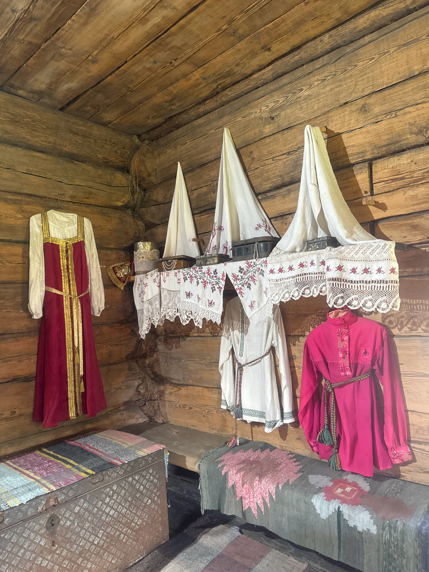 The image depicts the interior of a rustic wooden room, likely a museum exhibit, showcasing traditional clothing and furnishings. The walls and ceiling are constructed from rough-hewn logs, with visible joints and imperfections.
Hanging on the left wall is a long, sleeveless red dress with gold trim and a white long-sleeved undergarment. To its right, on a wooden shelf, are various items including what appears to be a decorative headpiece, several small ornate boxes, and intricately embroidered white textiles with floral patterns.
Below the shelf, two more traditional outfits are displayed. The one on the left is a white, long-sleeved tunic cinched with a braided belt. The one on the right is a bright pink, long-sleeved tunic also with a braided belt and embroidered details on the chest and cuffs.
In the foreground, two old, metallic chests are visible, each topped with colorful woven mats. A woven rug with geometric patterns lies on the floor in front of the chests. The overall impression is of a preserved historical setting.