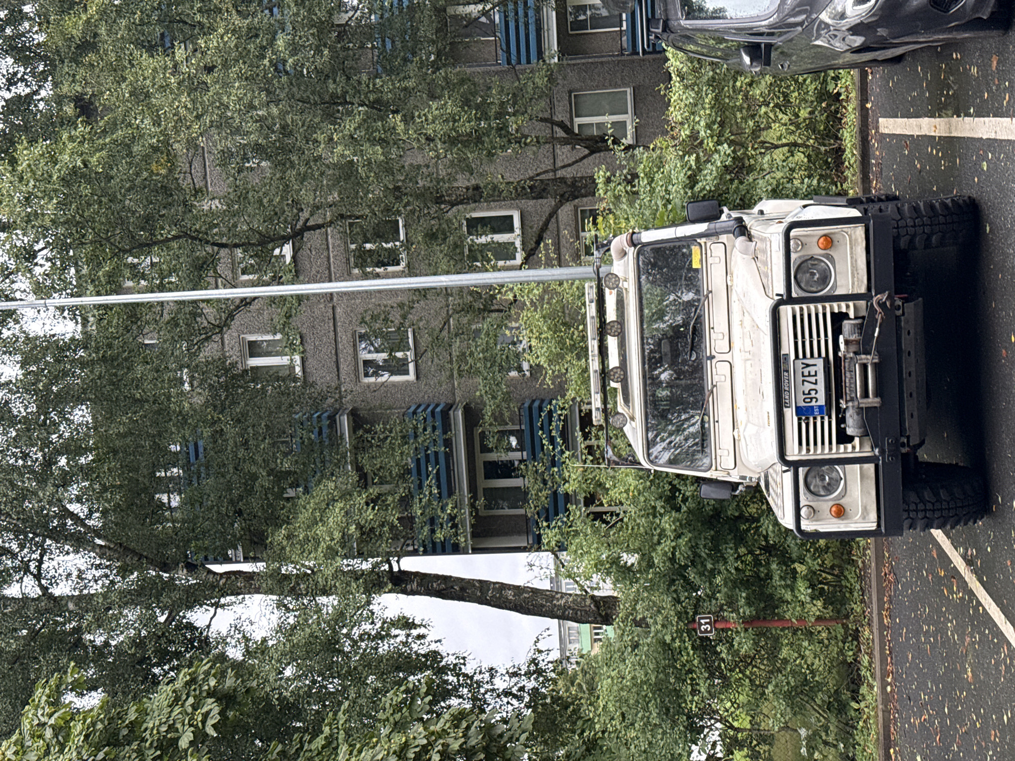 A light-colored Land Rover Defender is parked in an asphalt parking lot in front of a multi-story apartment building. The building is made of gray concrete with blue-railed balconies and white-framed windows. Trees with lush green foliage surround the building and the parking lot. A street sign with the number "31" is visible on a red post to the left of the Land Rover. The weather appears overcast, and fallen leaves are scattered on the wet ground. The Land Rover has a winch mounted on its front bumper and auxiliary lights on the roof. A dark-colored vehicle is parked to the right of the Land Rover. The license plate of the Land Rover reads "95 ZEY". The scene is set in the city of Tallinn, Estonia.