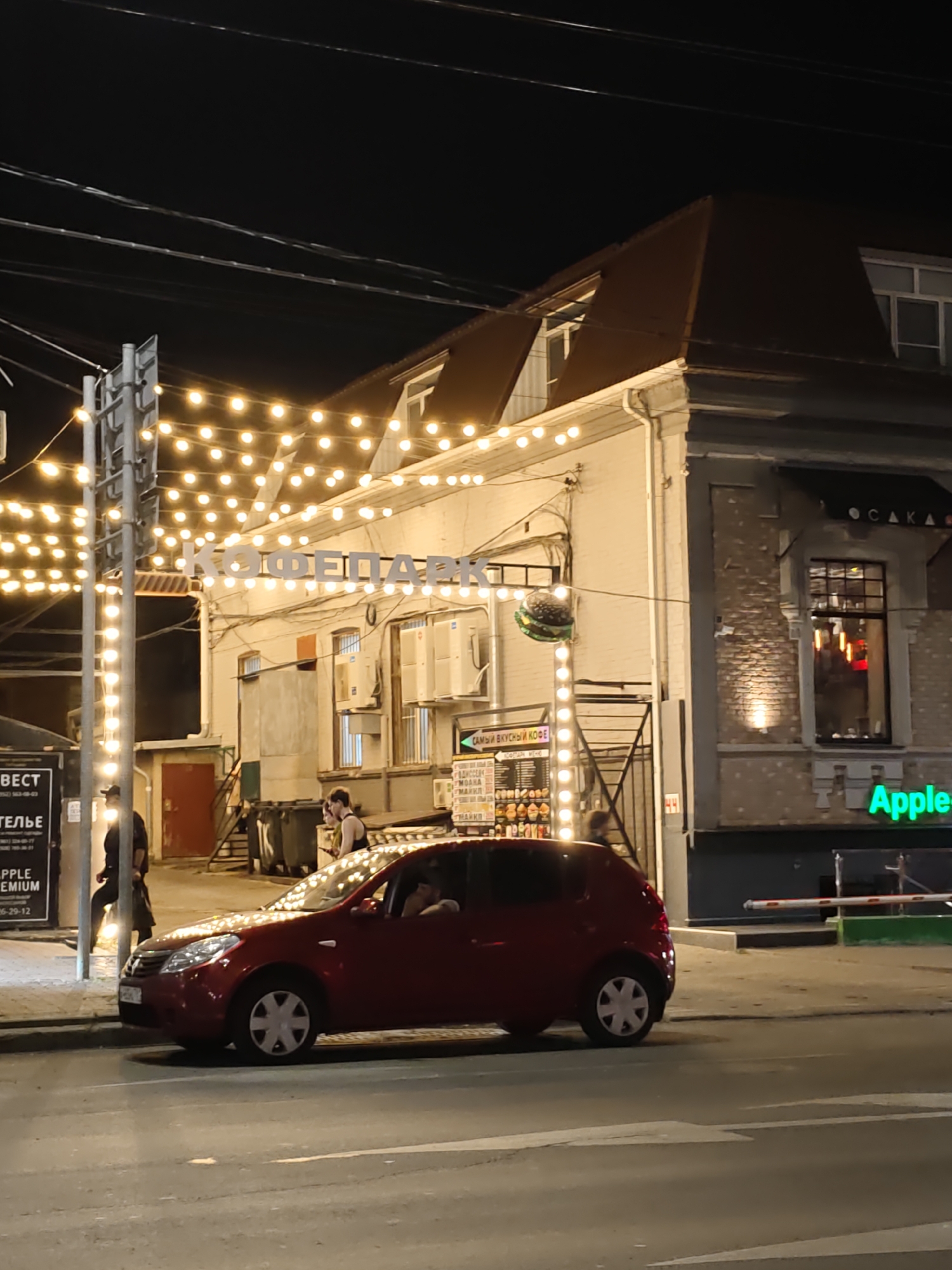 The image shows a street scene at night in Rostov-on-Don, Russia. The main subject is a red car parked on the side of the road. Above the car, a building is visible with lights strung along its facade and a sign that reads "КОФЕПАРК" (KofePark). To the right of the building, a green neon sign for "Apple" is illuminated. People are visible near the building, some standing and one walking. The street is paved, and there are streetlights and power lines overhead. The overall impression is of a commercial area at night, likely a cafe or restaurant.
