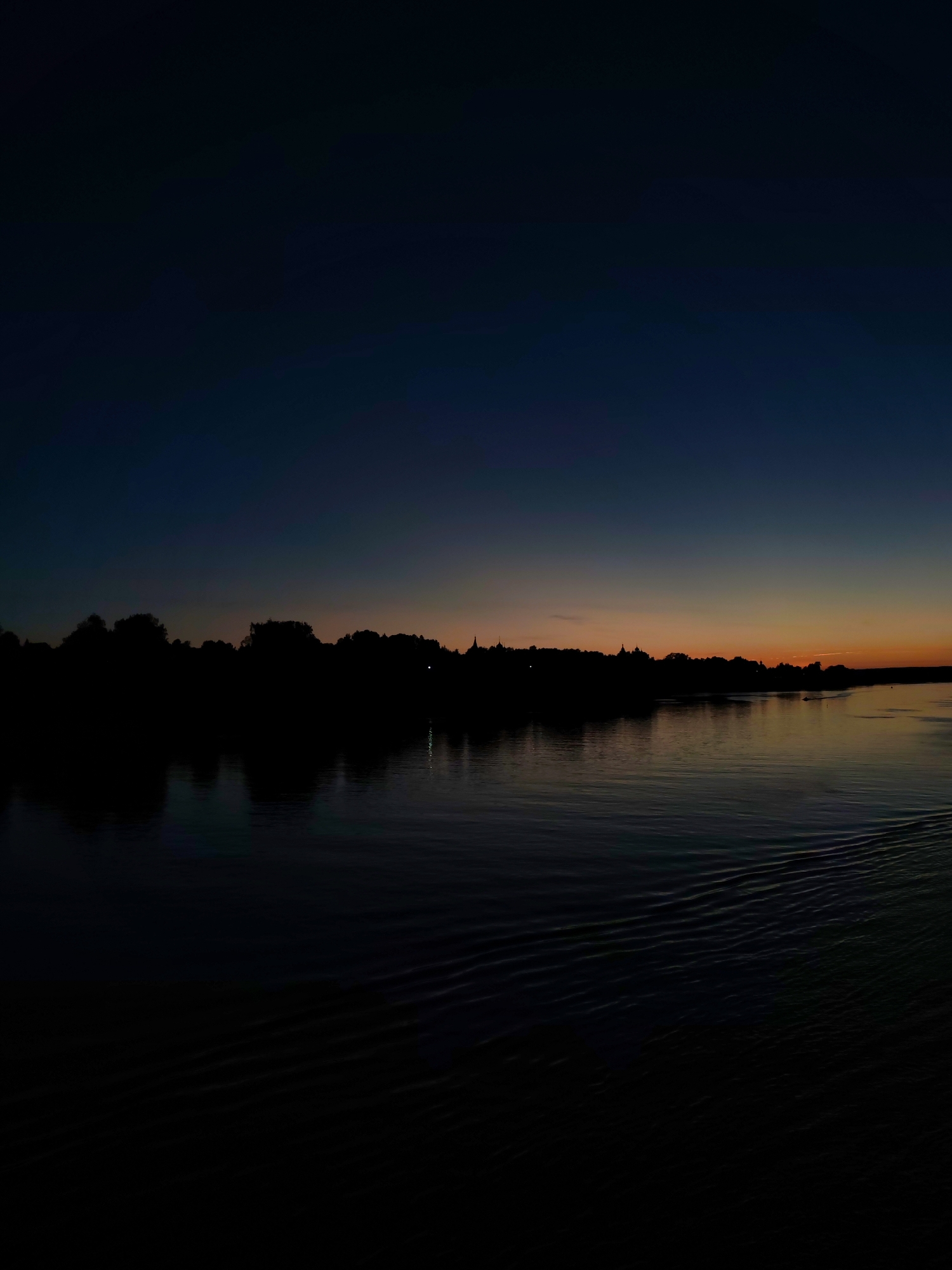A wide shot captures a calm body of water reflecting a dark, silhouetted shoreline under a twilight sky. The sky transitions from a deep navy blue at the top to a soft orange and yellow hue near the horizon, suggesting the aftermath of a sunset. The shoreline is densely populated with trees and buildings, all rendered in shadow, creating a stark contrast against the lighter sky. Small, indistinct lights are visible along the shoreline. The water in the foreground displays subtle ripples and wave patterns, hinting at movement, possibly from a boat, with a wake visible on the right side of the image. The overall scene is peaceful and atmospheric, with a muted color palette dominated by dark tones.