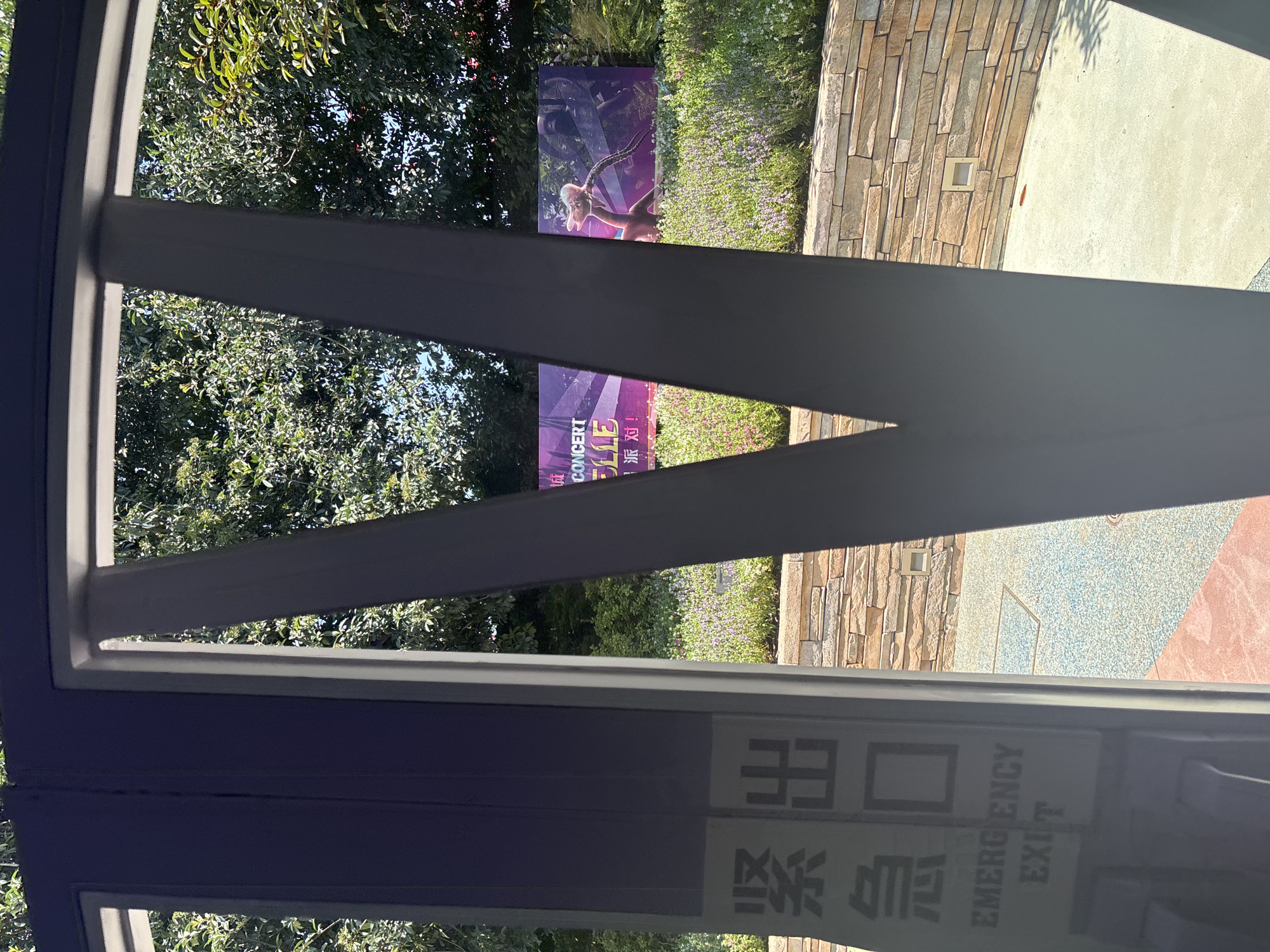 The image shows a view through a dark gray V-shaped structure, likely a doorway or partition, looking out onto an outdoor scene. On the left side of the foreground, a dark purple sign with white text is visible, which appears to be an emergency exit sign with Chinese characters followed by "EMERGENCY EXIT". In the background, a large banner with purple and pink hues is displayed horizontally, advertising a concert. The banner features imagery of a character with light-colored hair and a long neck, possibly from a performance or event. Below the banner, there is a wall constructed of stacked, light-brown stones, and beyond that, a paved ground with a speckled pattern of blue and pinkish-red. The background is filled with lush green trees and some flowering plants, suggesting an outdoor park or public space. The lighting indicates it is daytime with sunlight casting shadows. Based on the presence of Chinese characters on the emergency exit sign, it is plausible this location is in China. While the specific city is not explicitly stated, if this scene were to be located in Pudong, China, it would likely be a public area such as a park, entertainment venue, or shopping complex.
