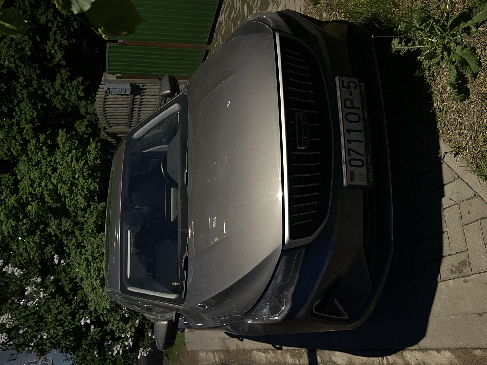The image shows the front of a dark-colored Geely car parked on a paved surface. The license plate reads "0711 OP-5" and indicates that the car is registered in Belarus. The location is likely Minsk, Belarus, based on the country code on the license plate. The car is next to a patch of grass and some bushes. A green fence is visible in the background. The image appears to be taken during daytime, with shadows cast on the ground.