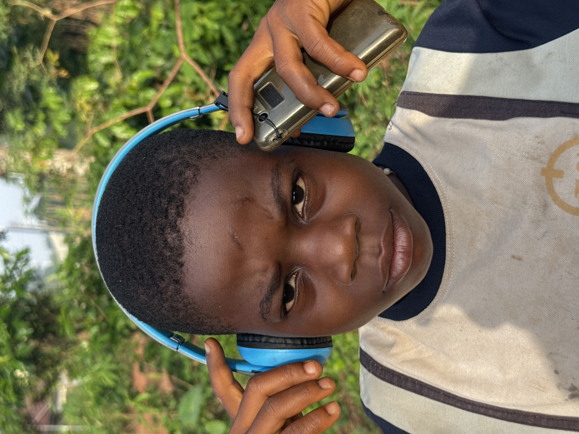 In Eziudo, Nigeria, an individual holds a gold-colored mobile phone against blue headphones worn on their head. The person wears a striped shirt with navy blue accents. The background consists of green foliage.