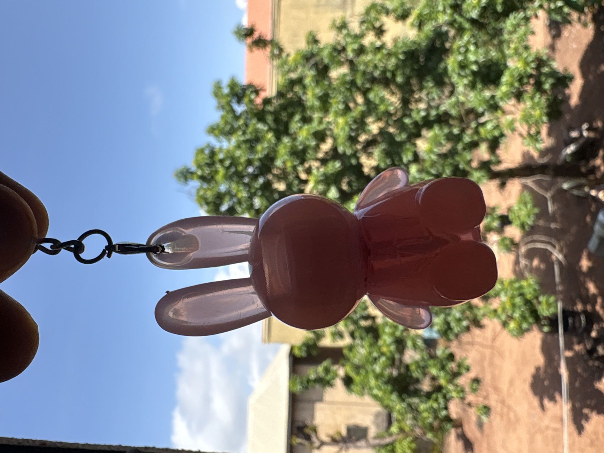 A translucent pink bunny-shaped keychain hangs from a metal chain, suspended against a backdrop of blue sky with scattered white clouds. In the background, blurred green trees with some visible brown branches are in focus. Behind the trees, parts of buildings are visible: a light-colored structure with a cream-colored roof on the left, and a yellow building with a reddish-orange roof on the right. The scene appears to be outdoors, possibly in a residential or urban area. This image was taken in Onitsha, Nigeria.