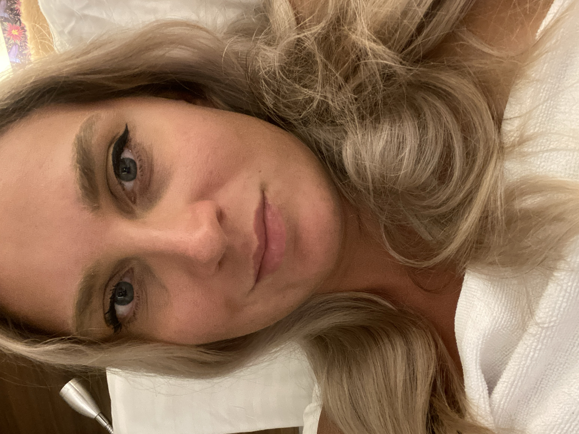 A close-up, indoor, horizontal selfie shows a woman lying in bed. She has long, blonde, wavy hair and fair skin. Her eyes are blue-green, and she is wearing winged eyeliner. Her lips are parted slightly, and she appears to be smiling faintly. She is wrapped in a white towel or blanket. The background is out of focus, but suggests a bedroom setting with a pillow and a bedside lamp. No specific location in Aksu, Turkey is identifiable.