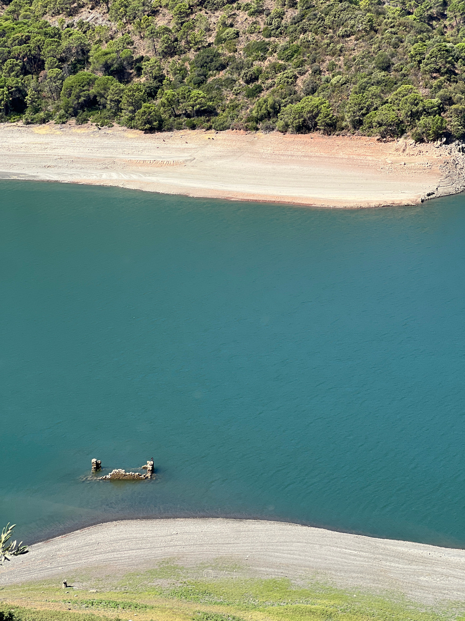 The image depicts a reservoir with a strip of dry land exposed along the shoreline. The water is a deep teal color, with ripples indicating a slight breeze. In the distance, a forested hillside rises, with a mix of green trees and exposed brown earth. Closer to the water, the shoreline is a sandy, light brown color, with visible erosion patterns. Partially submerged in the water, near the center of the frame, are the remains of what appears to be a stone structure, possibly a boat or building foundation. The overall scene suggests a dry season or a lowered water level in the reservoir. The location is the reservoir of the city of Istán, Spain.