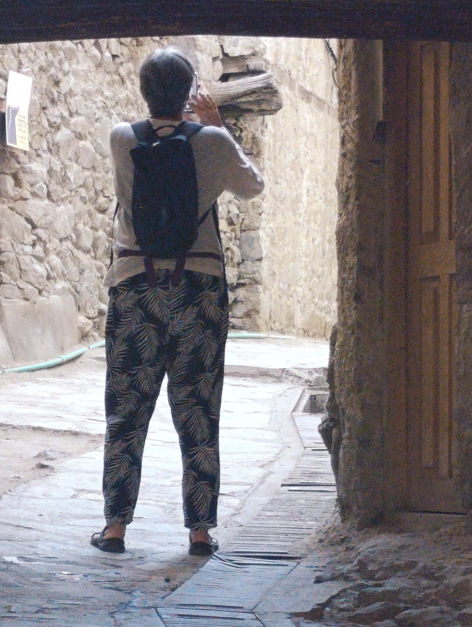 A person with gray hair, seen from behind, wearing a light-colored long-sleeved shirt, a dark backpack, and patterned pants, stands in a narrow alleyway. The person is holding a smartphone in their right hand, appearing to take a photograph. The alley is constructed of stone walls on both sides. To the left, a light-colored sign with "CCTV" written on it is visible on the wall. The ground is paved with stones and wooden planks. The scene is set in Leh, India, likely within the old city or a similar historical area characterized by its traditional architecture and narrow pathways.