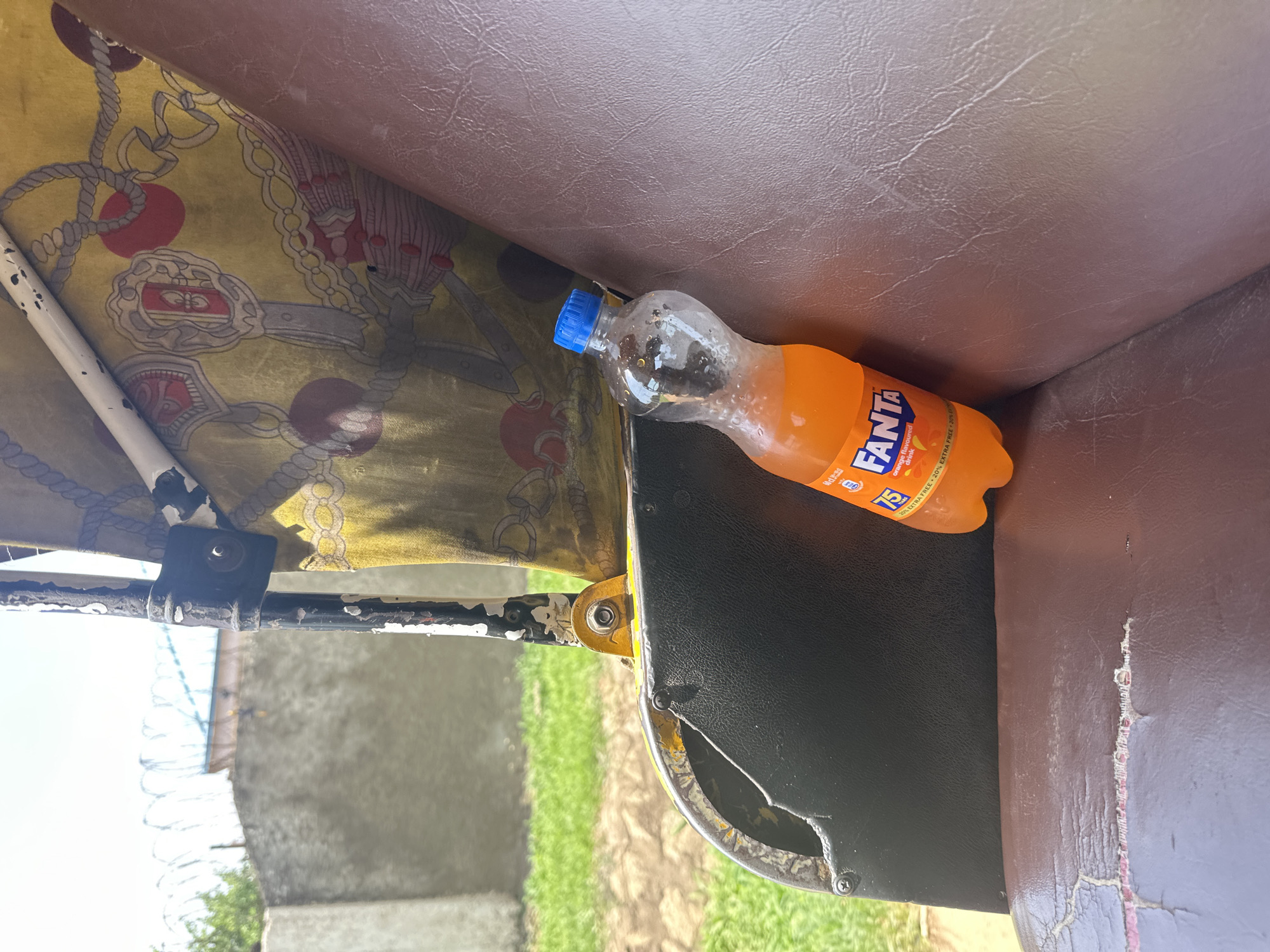 The image shows a close-up, high-angle view of an orange Fanta bottle with a blue cap, resting on the brown leather seat of what appears to be a rickshaw or tuk-tuk. The bottle is partially full, and some dark sediment is visible inside. The seat has a patterned yellow fabric underneath it, featuring a chain and crest design. The rickshaw's metal frame, painted white and black, is visible on the left side, along with a black metal clamp. In the background, out of focus, are a grey wall with barbed wire on top, green grass, and a bright sky, suggesting an outdoor setting during the daytime. The Fanta bottle's label is partially visible, displaying text that includes "FANTA," "orange flavoured drink," and "20% EXTRA FREE." The overall impression is one of casual transport in a warm, possibly tropical, environment.