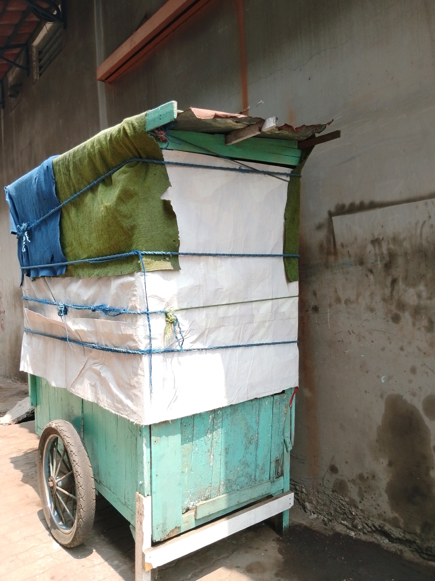 An abandoned, weathered, teal-colored wooden cart with a damaged tin roof sits near a rough, stained concrete wall in Pasuruan, Indonesia. The cart is partially covered with torn white and green tarps, secured by blue rope. A single, spoked wheel is attached to the front left side. The ground is paved with rough concrete.